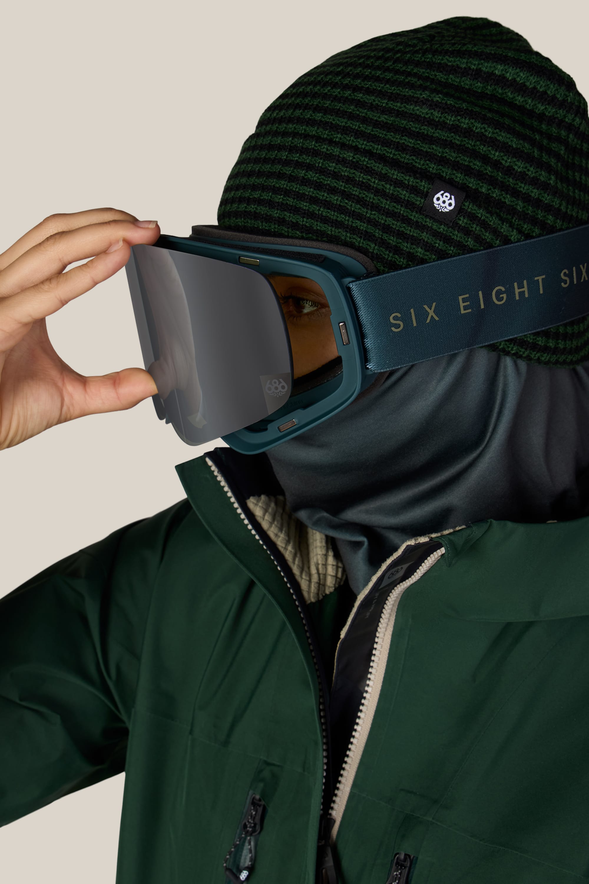 A person in a green striped beanie, face covering, and green jacket adjusts a pair of 686 SMARTY® Magnetic Goggles with “686” branding on the strap, set against a plain background.