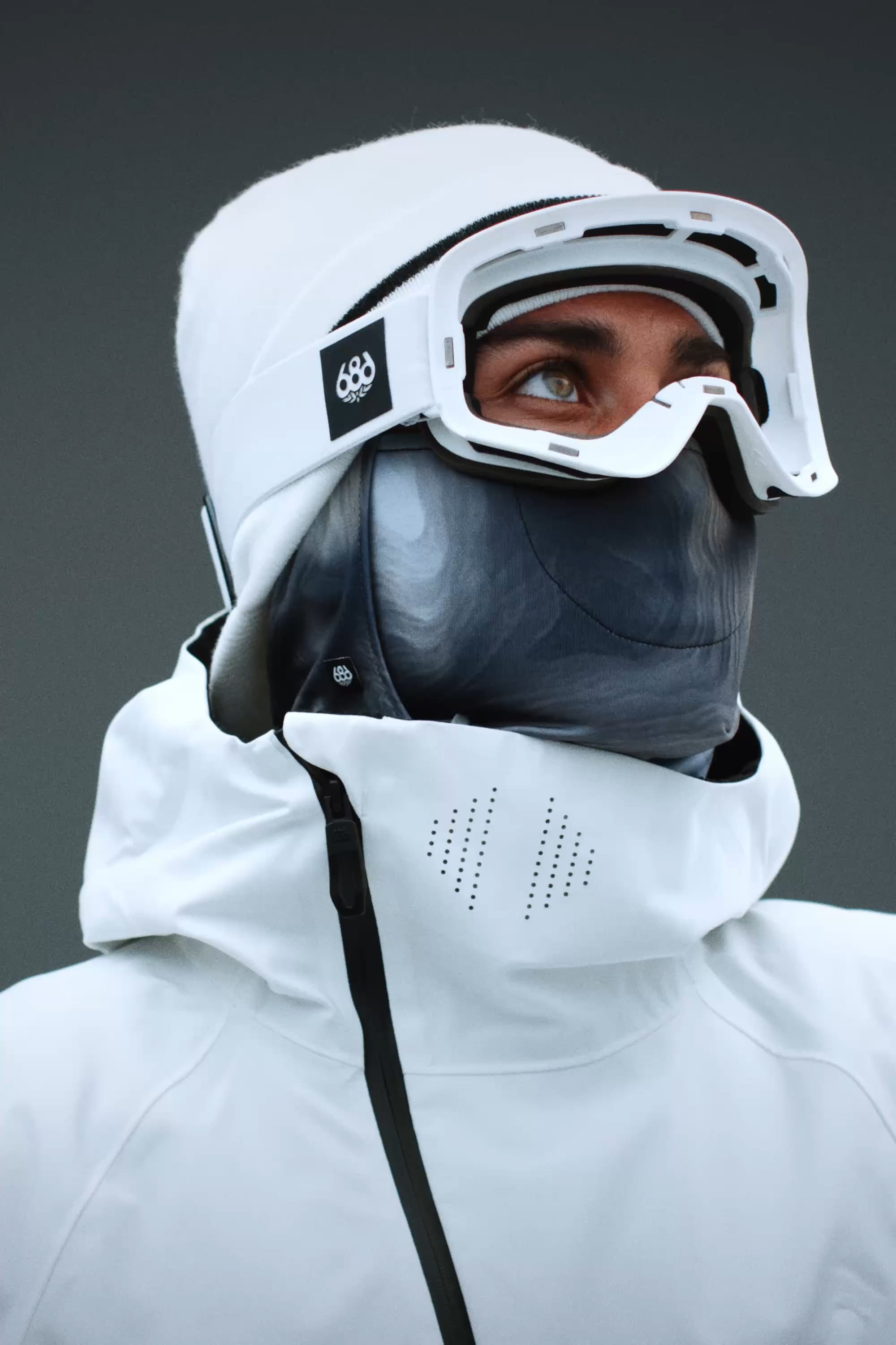 Wearing the 686 SMARTY® Magnetic Goggle by 686, a person dressed in white winter gear with a jacket and face mask looks upward against a plain gray background.