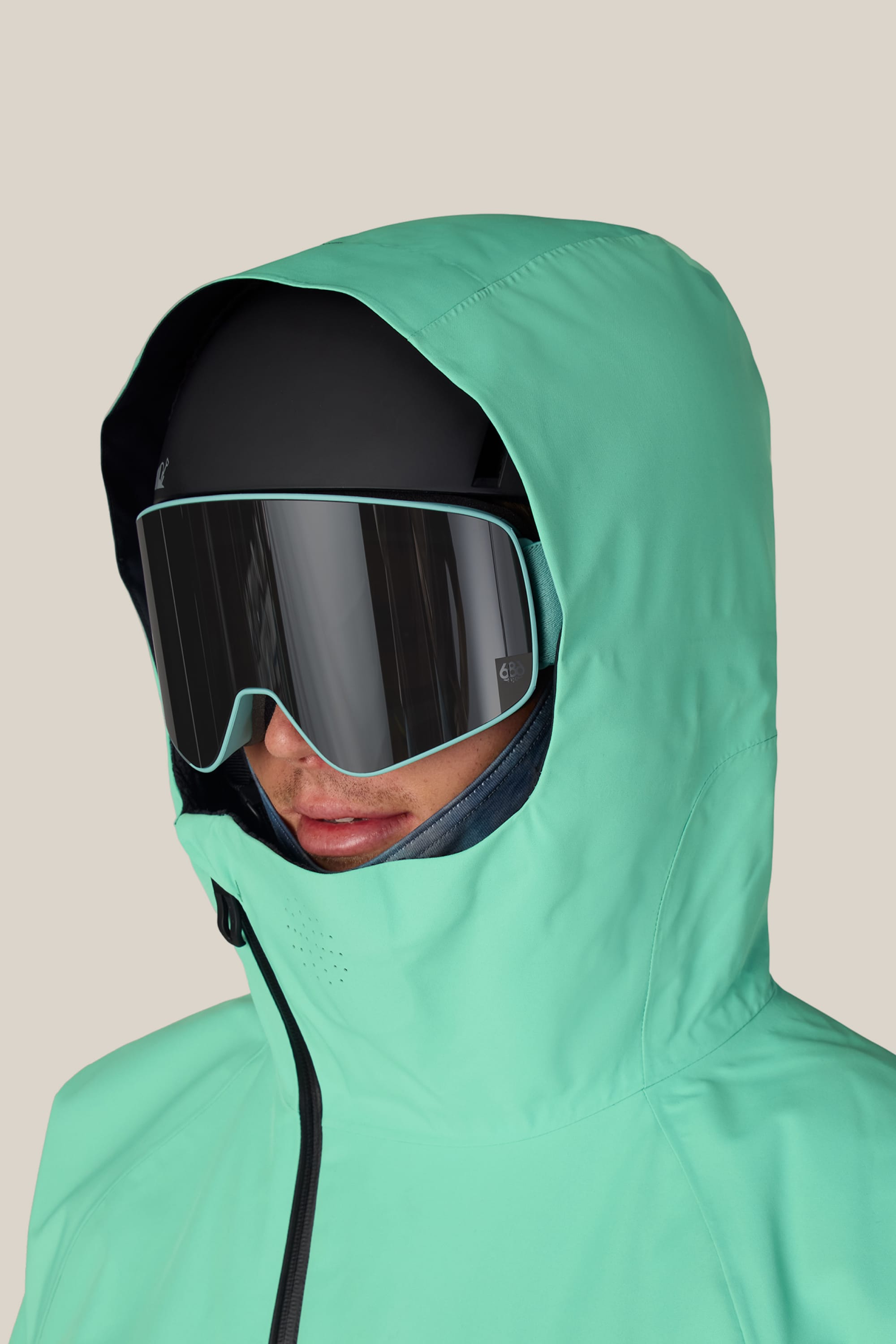 A person wearing a mint green hooded jacket, black ski helmet, and large 686 SMARTY® Magnetic Goggles by 686, set against a plain beige background.
