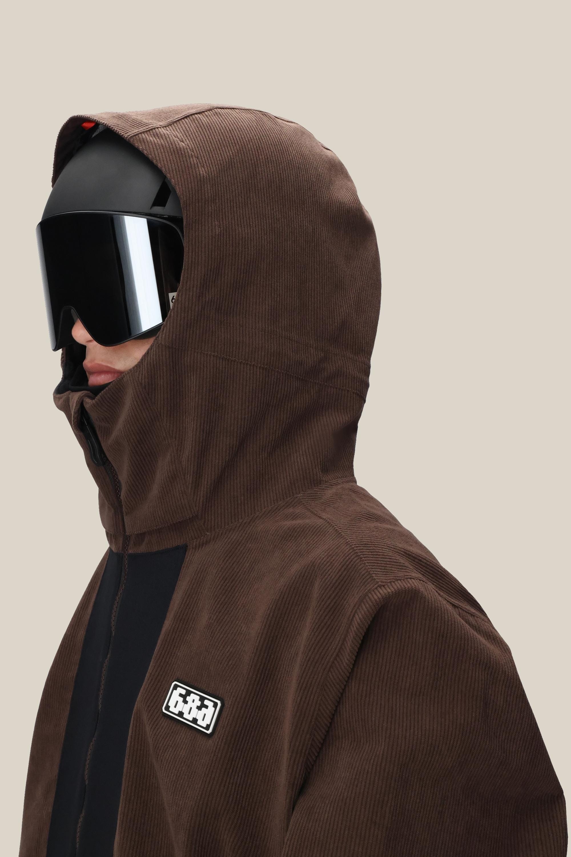 A person wears the 686 Men's Ghost™ 2.5L Anorak with the hood up, paired with black ski goggles and a black helmet. A patch with white text is visible on this rugged brown corduroy jacket from 686.