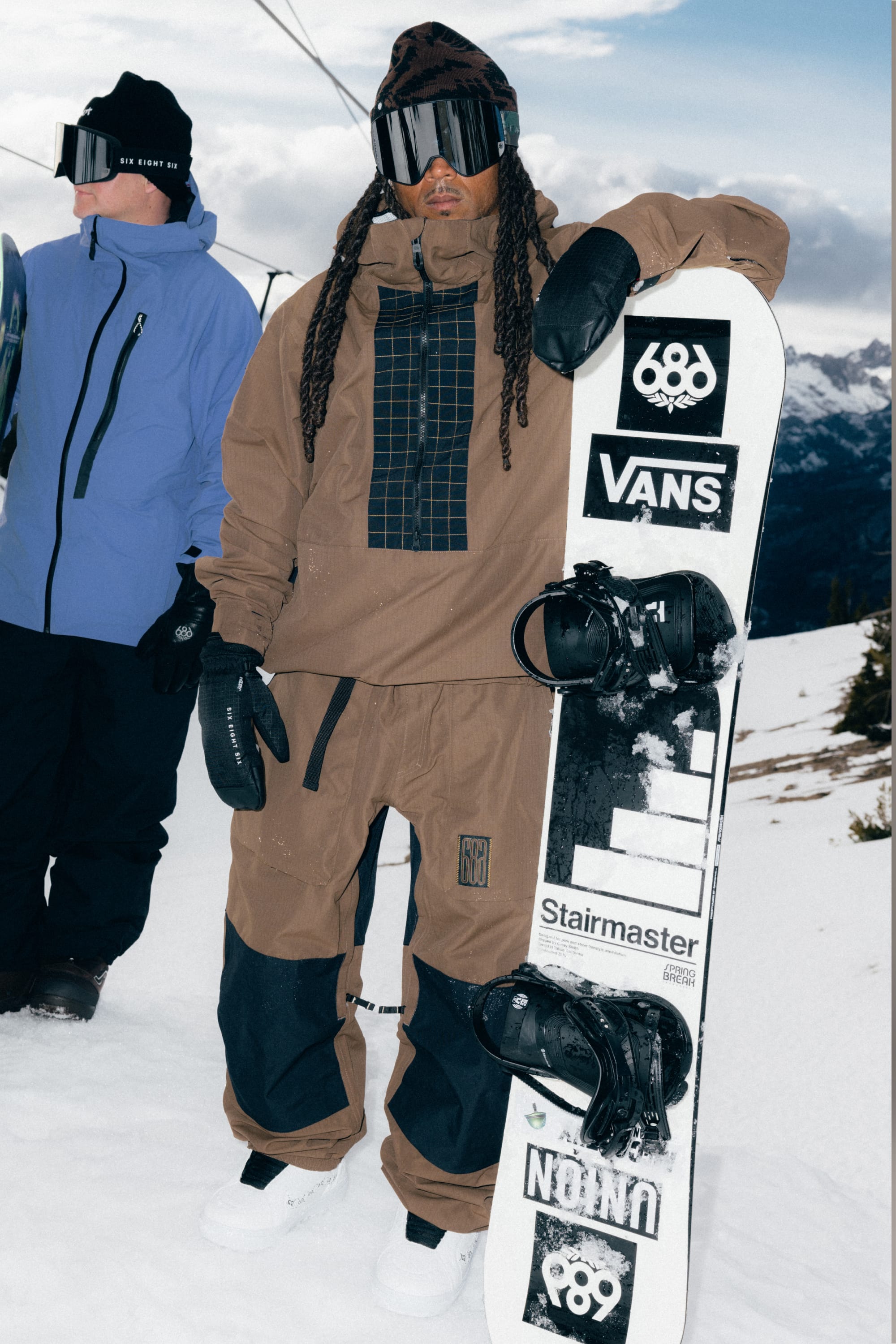 A person wearing the 686 Men’s Ghost™ 2.5L Anorak stands on a snowy slope in brown and black snow gear with goggles, holding a white snowboard decorated with stickers. Another person in a blue jacket appears in the background, with snowy mountains and trees visible behind them.