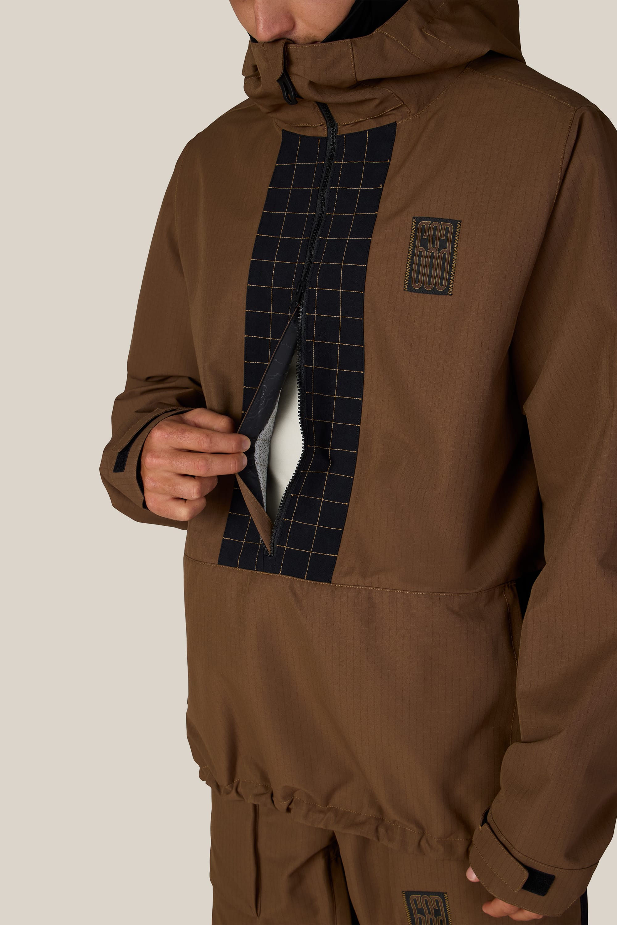 A person wearing the 686 Men’s Ghost™ 2.5L Anorak from 686, featuring a brown color with a black grid pattern, unzips the front flap to reveal a white inner lining. The anorak has a high collar and a matching patch on the chest, with only the torso and lower face visible.