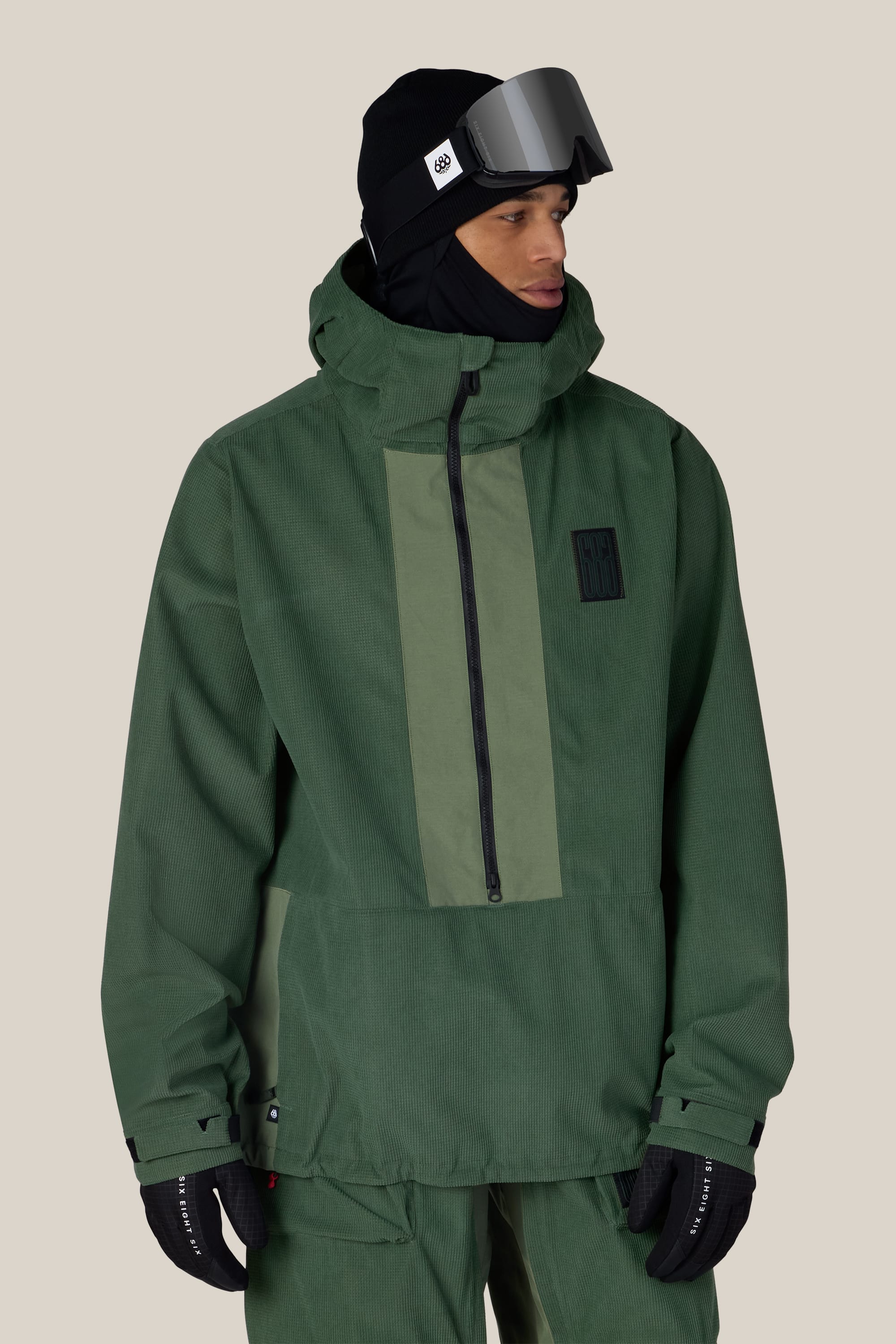 A person wearing the 686 Men’s Ghost™ 2.5L Anorak by 686 in green, paired with matching pants, black gloves, a black beanie, and ski goggles, stands against a plain beige background.