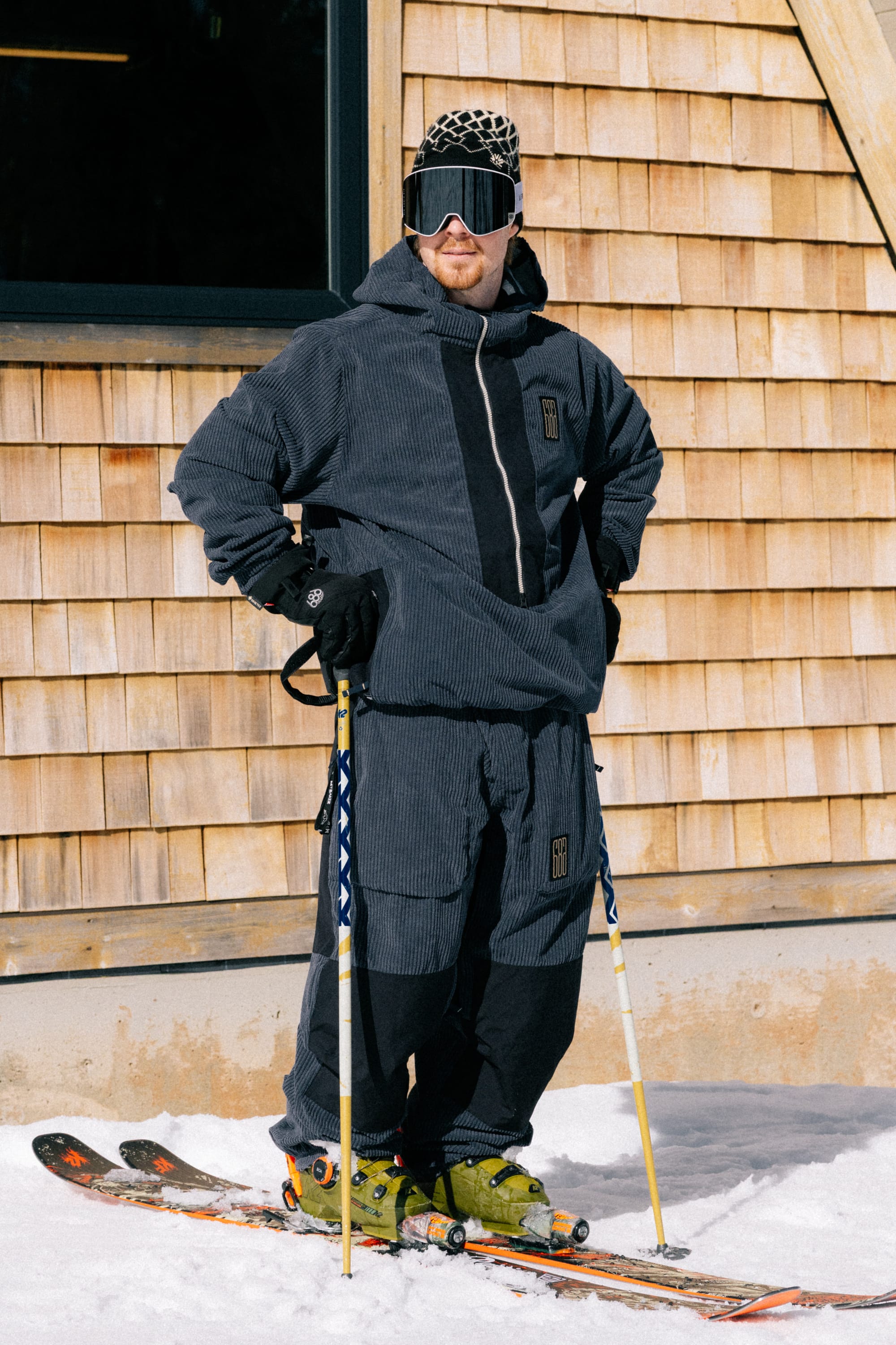 Wearing the 686 Men’s Ghost™ 2.5L Anorak, a skier stands on snowy ground with skis and poles, ski goggles, and a hat in front of a wooden building.