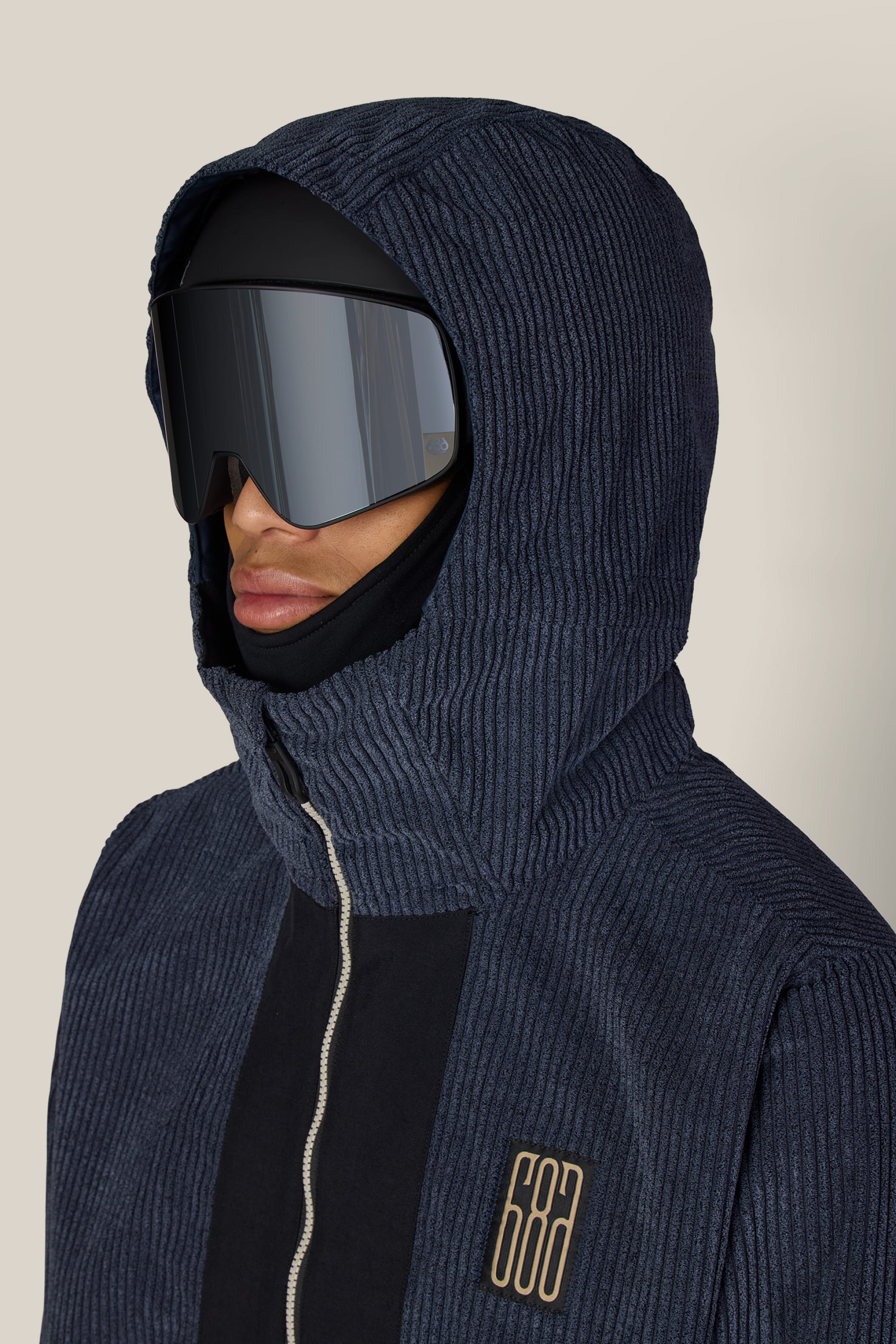 A person wearing the 686 Men’s Ghost™ 2.5L Anorak—a textured navy hooded jacket with a beige 686 logo on the chest—paired with black ski goggles and a black balaclava. The background is plain and light-colored.