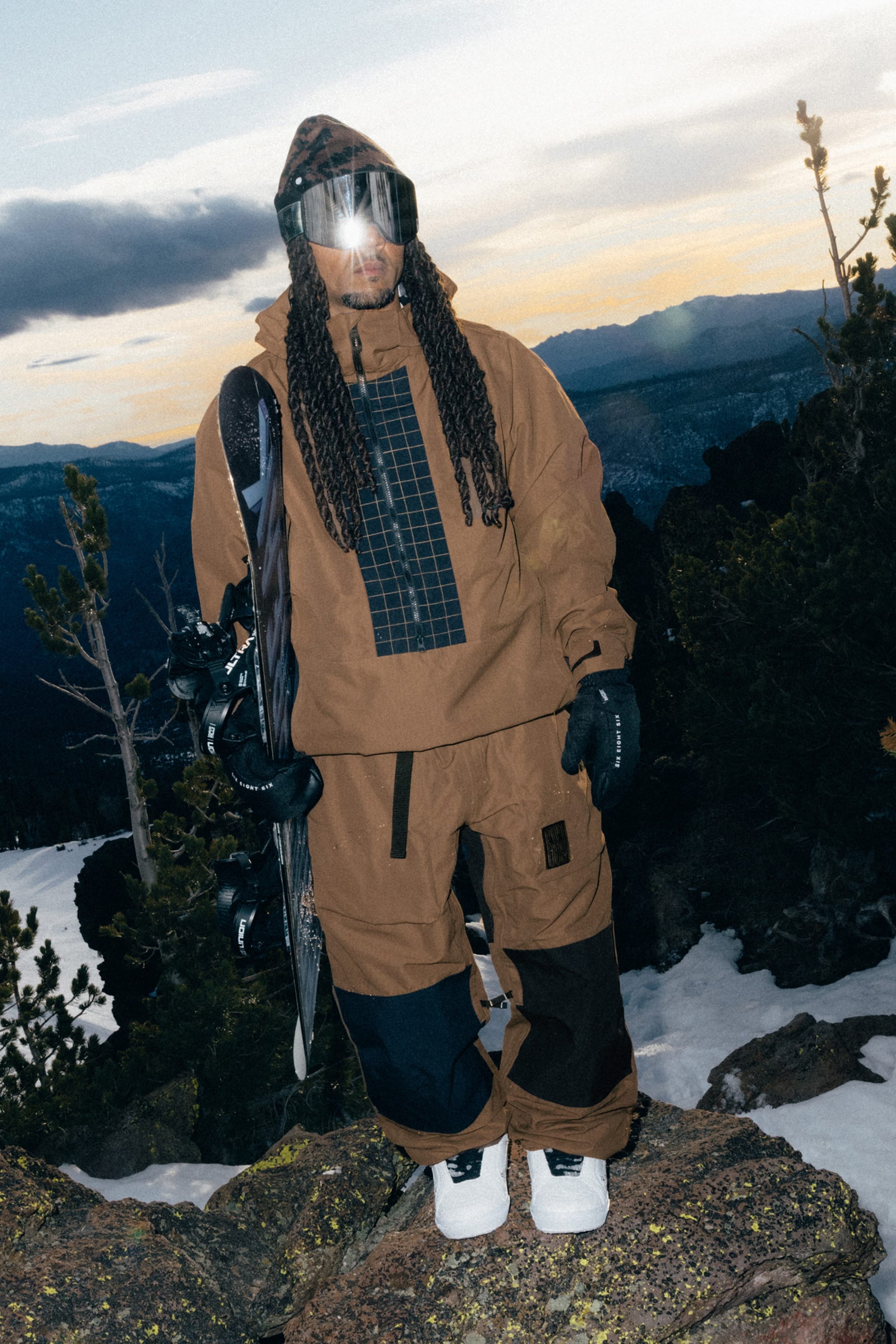 Wearing the 686 Ghost™ 2.5L Pant by 686, a person stands on a rocky, snowy mountain in brown snow gear and reflective goggles, holding a snowboard with trees and a scenic, cloudy sky in the background.