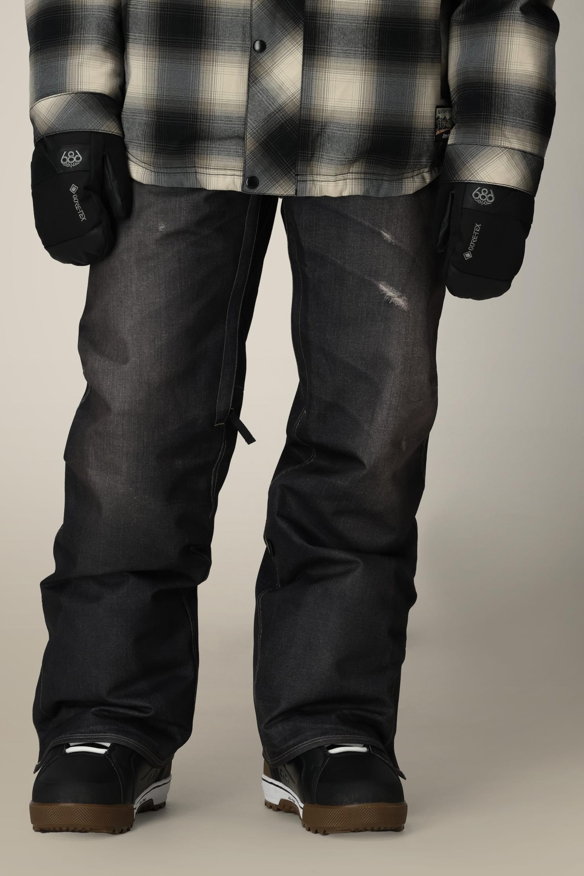 A person wearing a black and white plaid shirt, black snowboard gloves, 686 Men's Deconstructed™ Denim Pant by 686, and black snowboard boots stands against a neutral background.