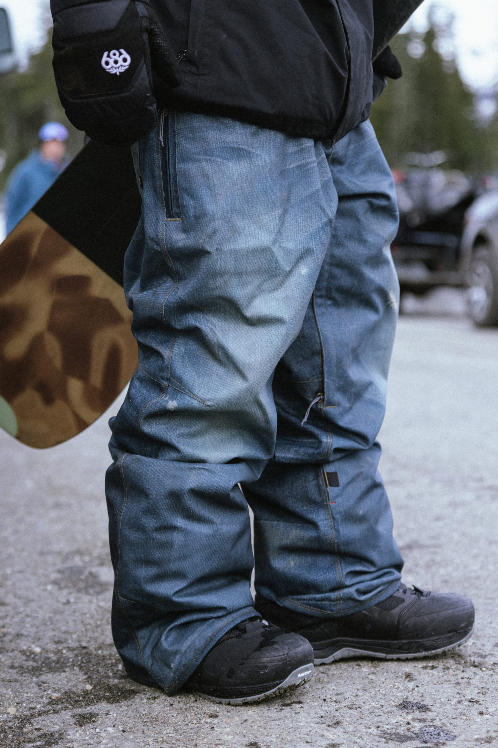 A person in 686 Men's Deconstructed™ Denim Pant, black snow boots, and a black jacket stands on snowy pavement holding a camouflage snowboard, with blurred people and trees in the background.