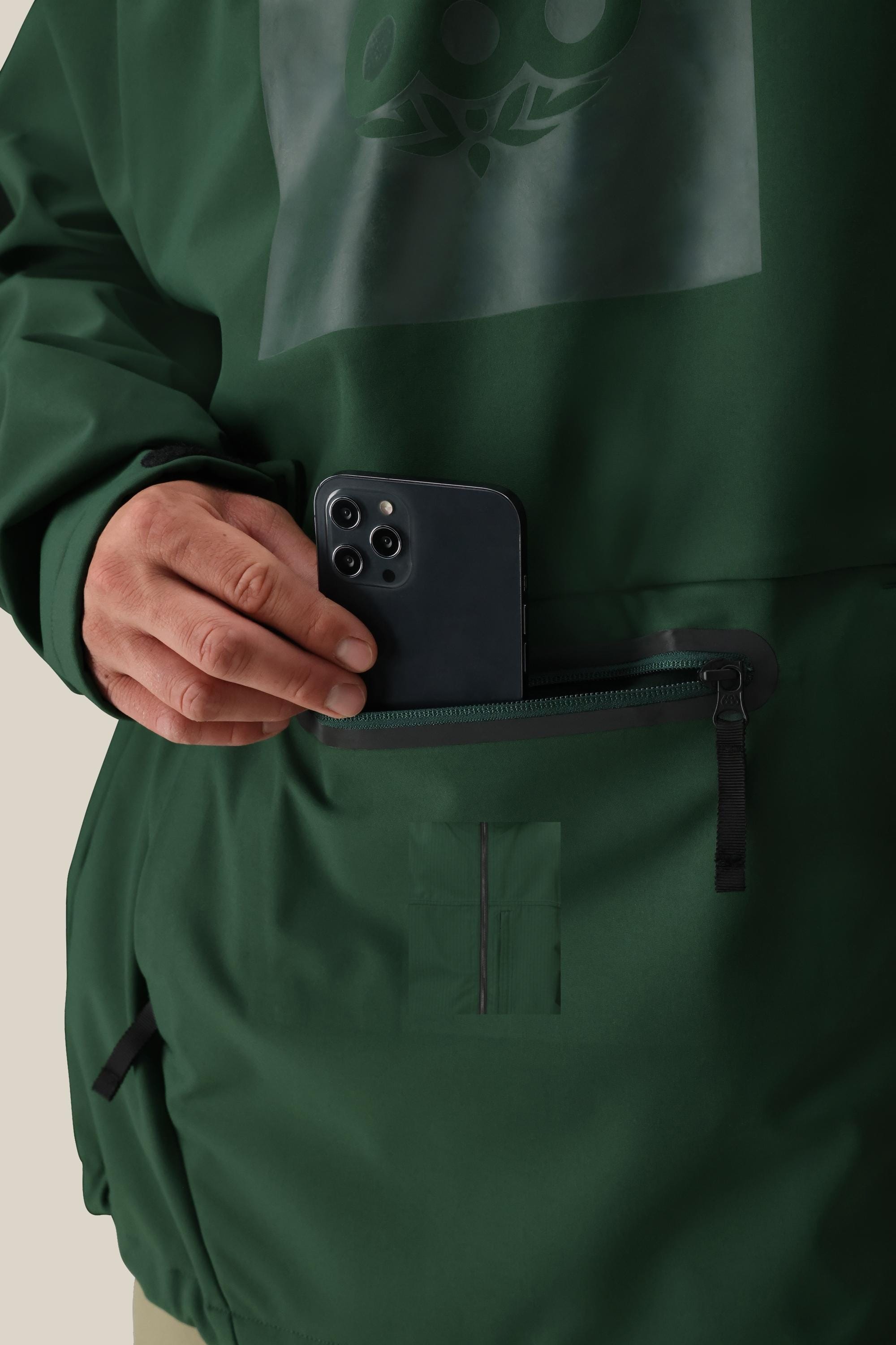 A person wearing the 686 Men’s Waterproof Hoody places a black smartphone into a front zippered pocket on the hoody. Only their hand and part of their body are visible.