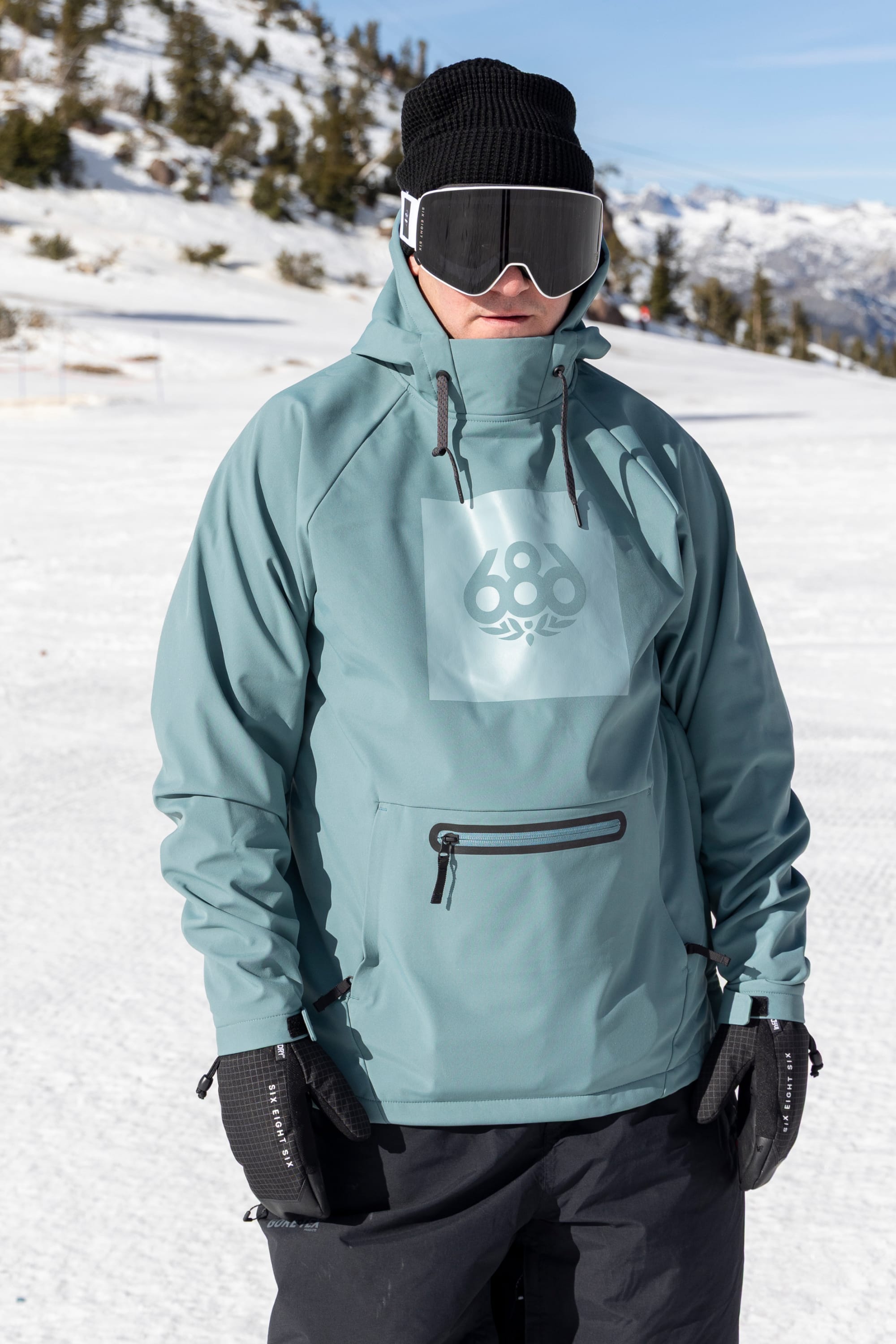 A person wearing the 686 Men’s Waterproof Hoody by 686, along with a black beanie, gloves, and ski goggles, stands on a snowy mountain slope with trees and mountains in the background under a clear blue sky.