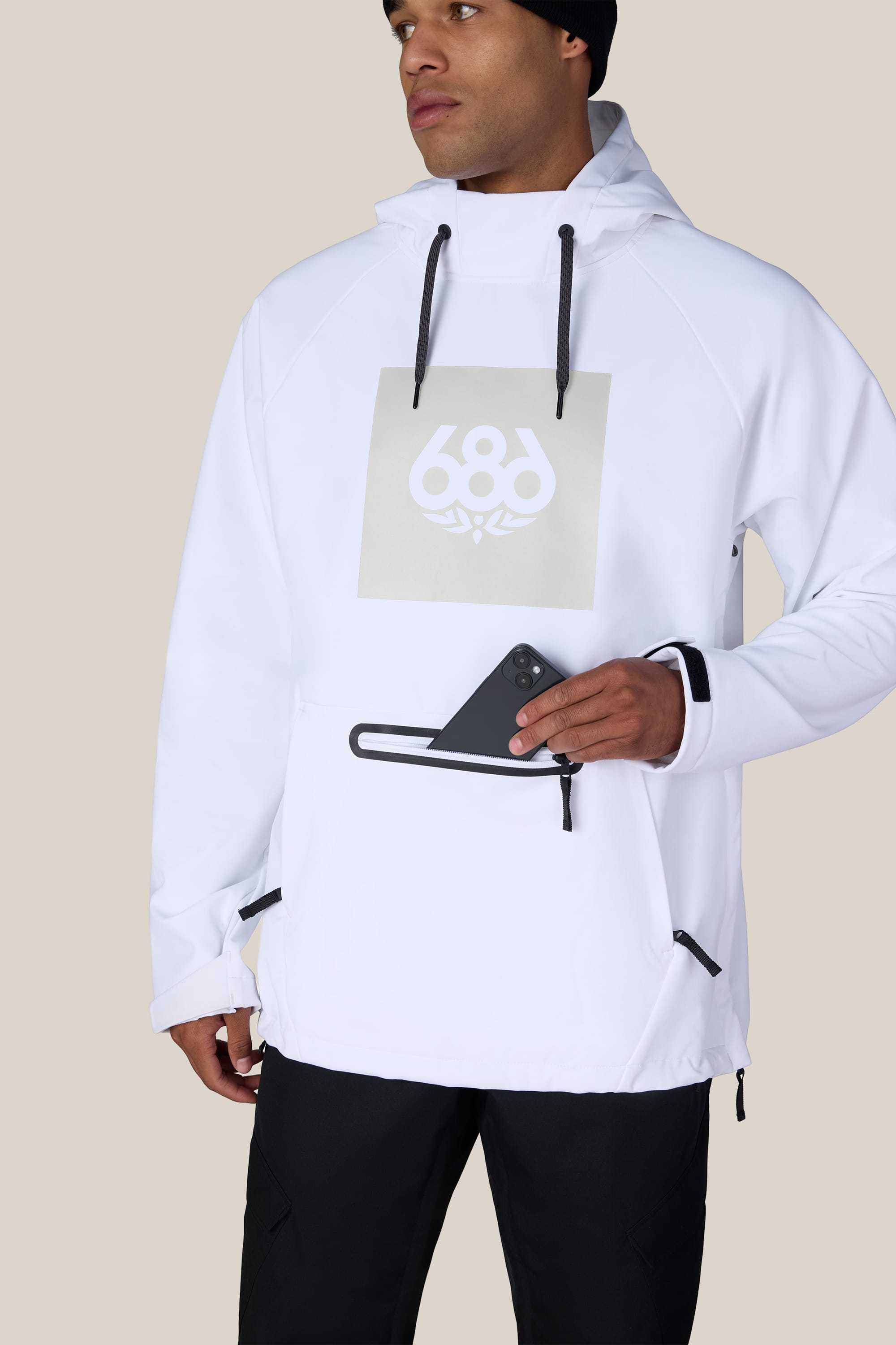 A person wearing the 686 Men’s Waterproof Hoody in white, paired with black pants and a black beanie, places a smartphone into the hoody’s zippered front pocket. The background is beige.