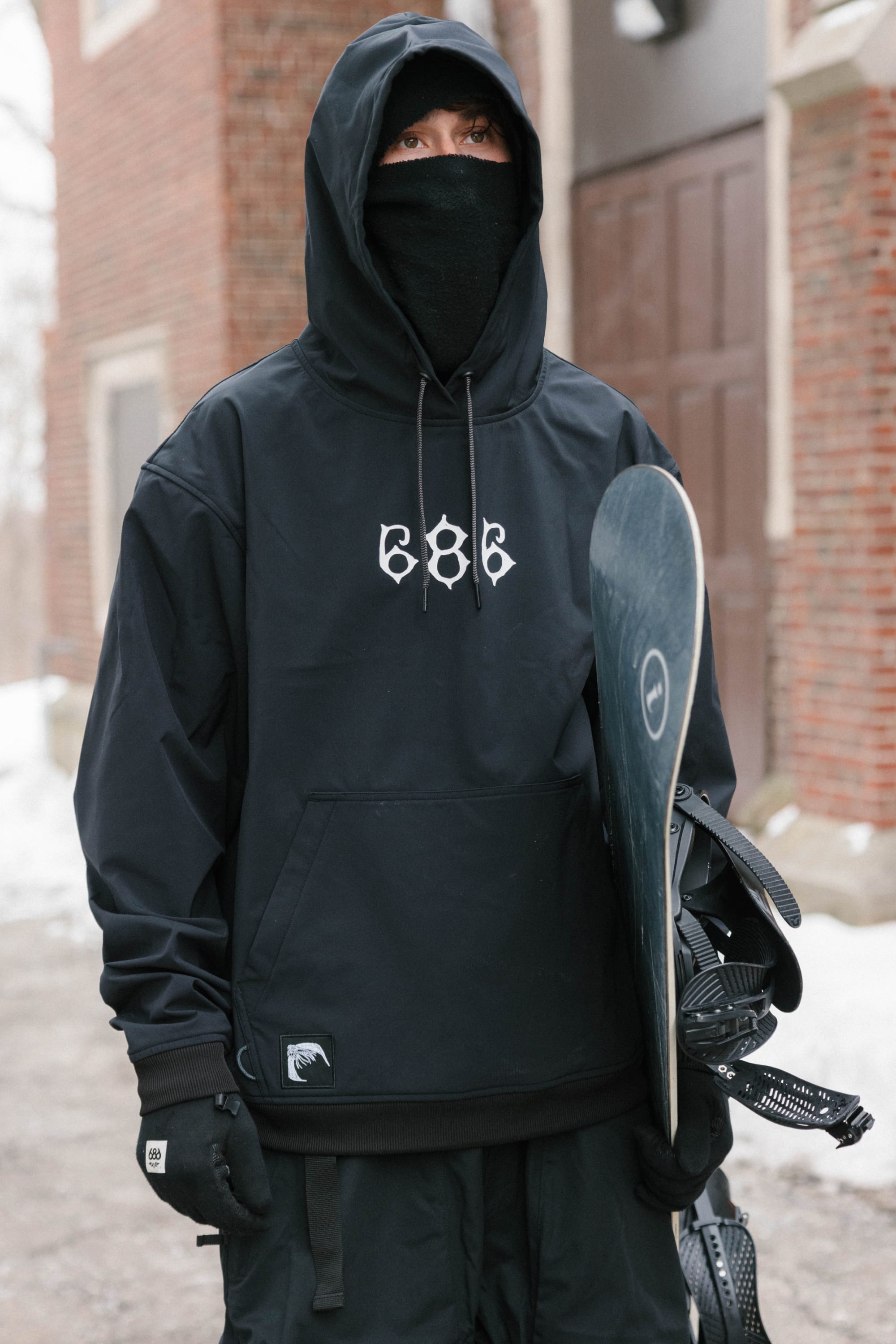 Wearing a 686 Mens Waterproof Team Hoody, black gloves, and a face mask, a person holds a snowboard while standing outside near a brick building with snow on the ground.