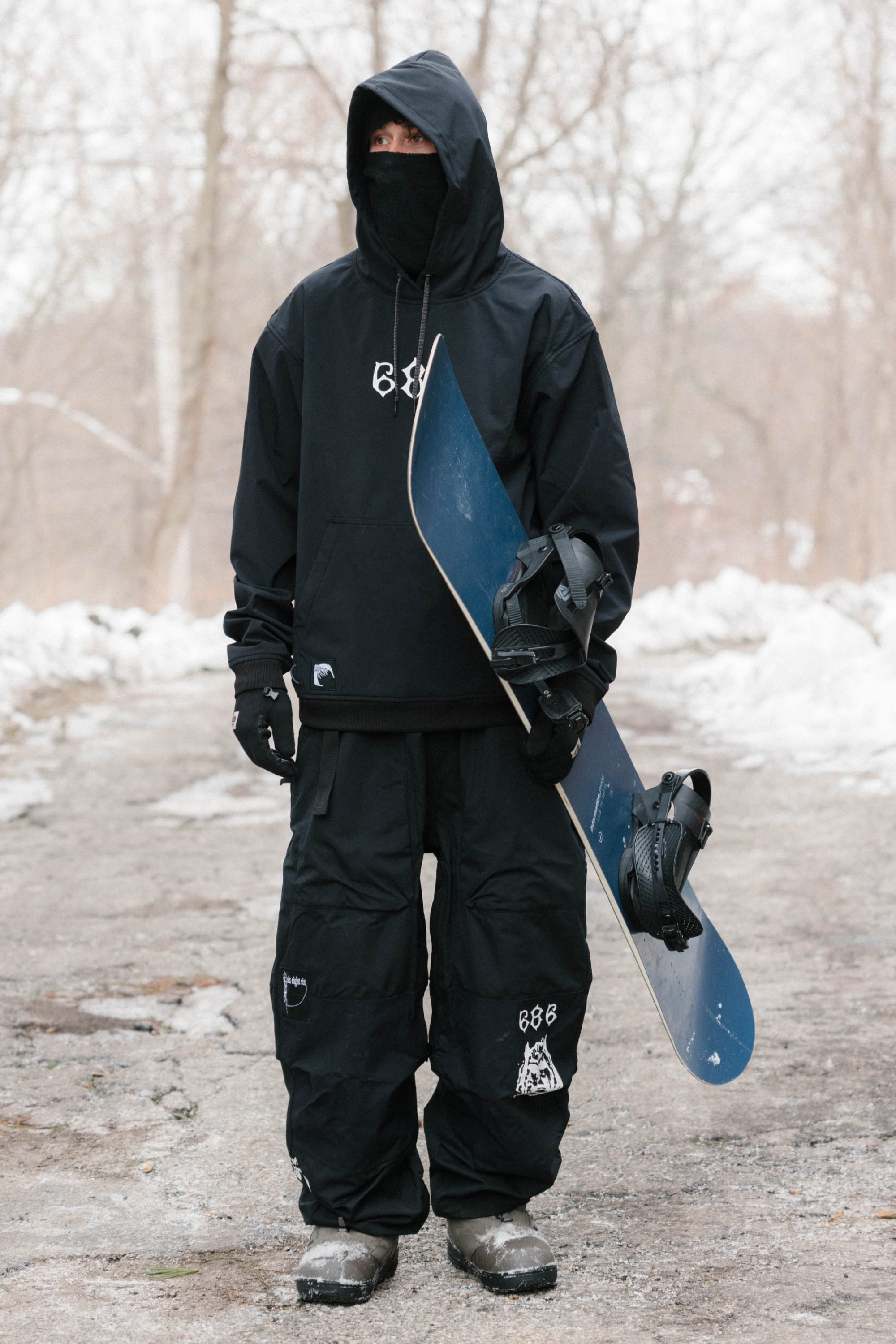 Dressed in the 686 Mens Waterproof Team Hoody, a person stands on a snowy path holding a snowboard vertically, with bare trees and snow in the background.
