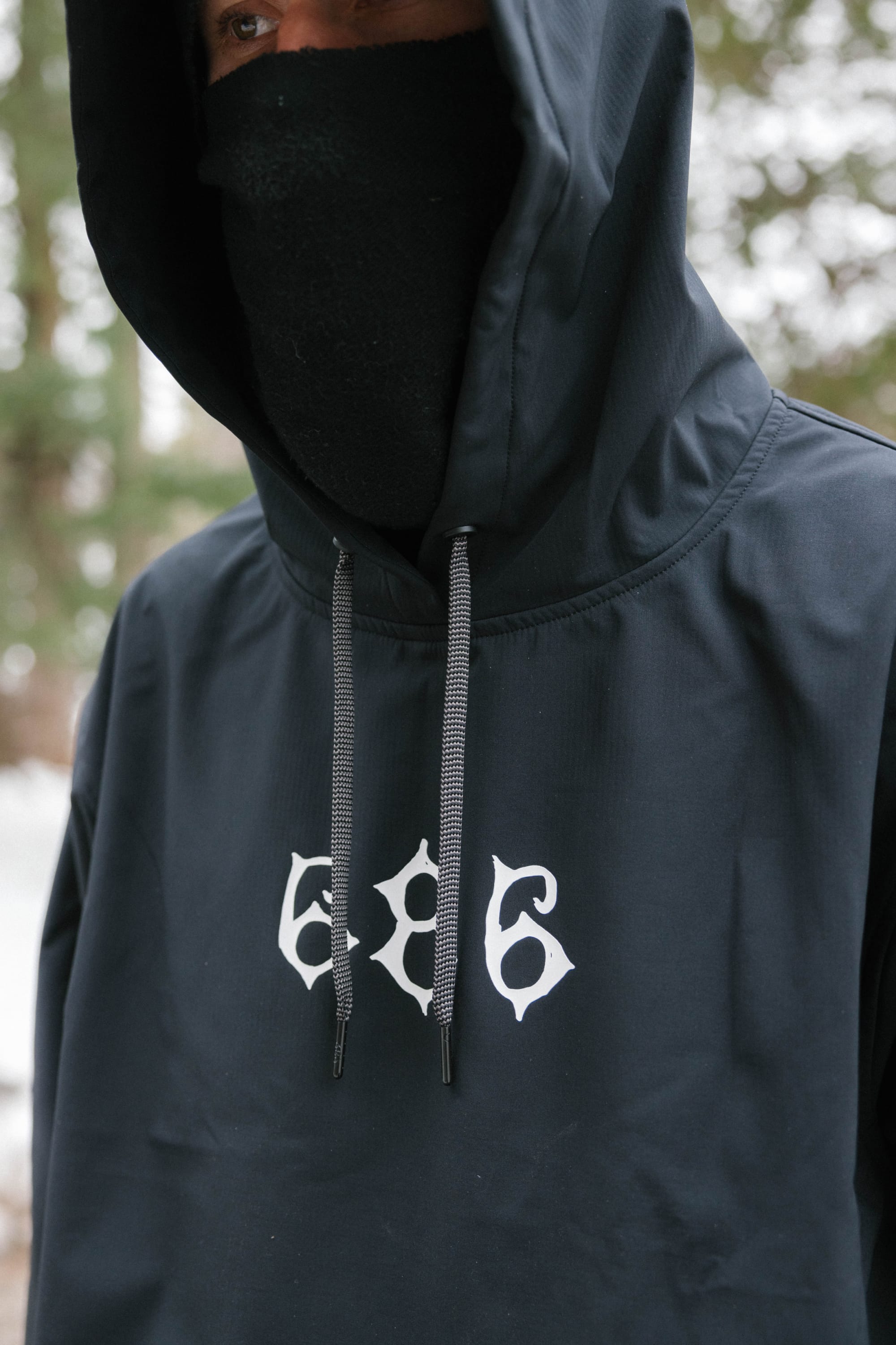 A person wears the 686 Mens Waterproof Team Hoody in black with “686” printed in white on the front, paired with a black mask. Blurred trees are visible in the background.