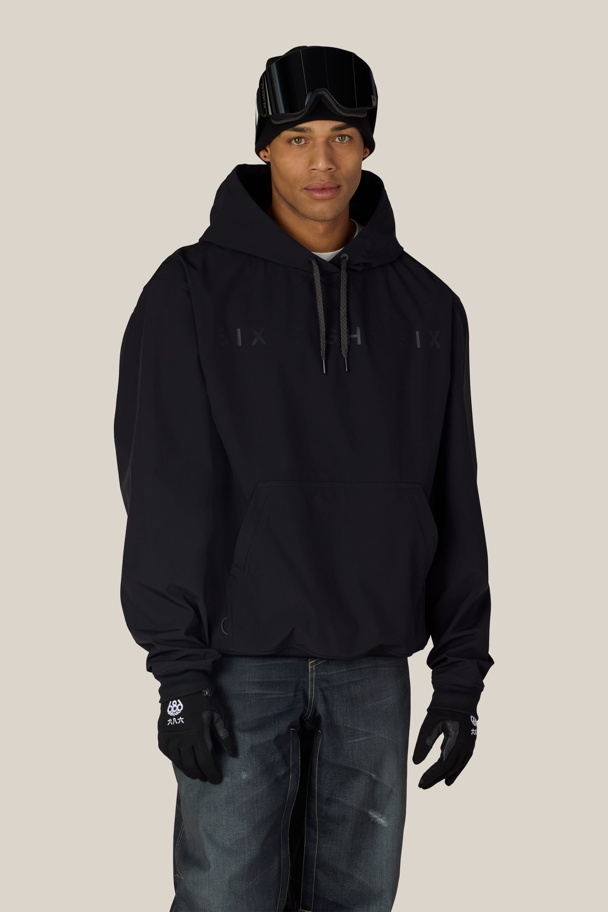 A man wearing the 686 Men’s Waterproof Team Hoody from 686, paired with black gloves, dark jeans, and ski goggles on his forehead, stands against a plain light background and looks at the camera with a neutral expression.