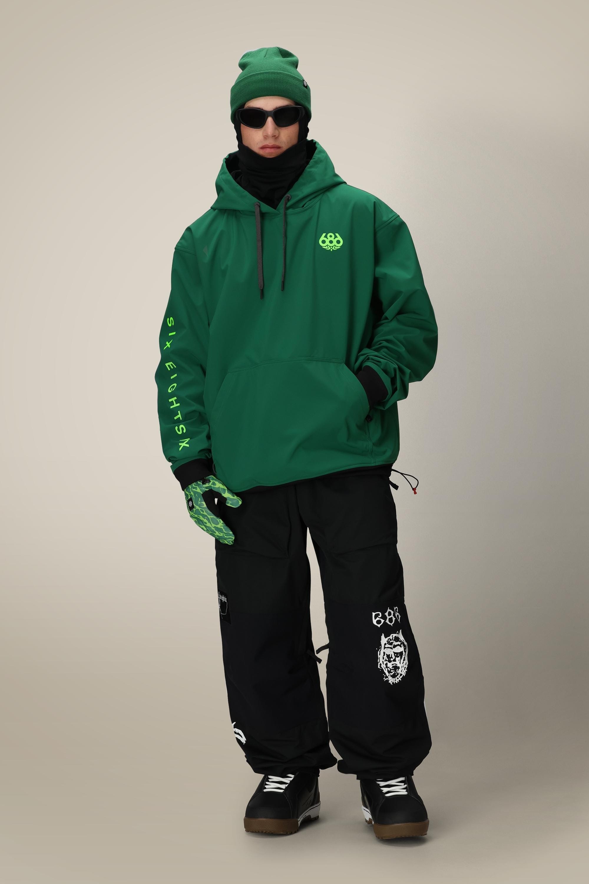 A person wearing the 686 Mens Waterproof Team Hoody, a green beanie, sunglasses, black scarf, patterned green gloves, and baggy black pants with white graphic designs stands against a plain light background.