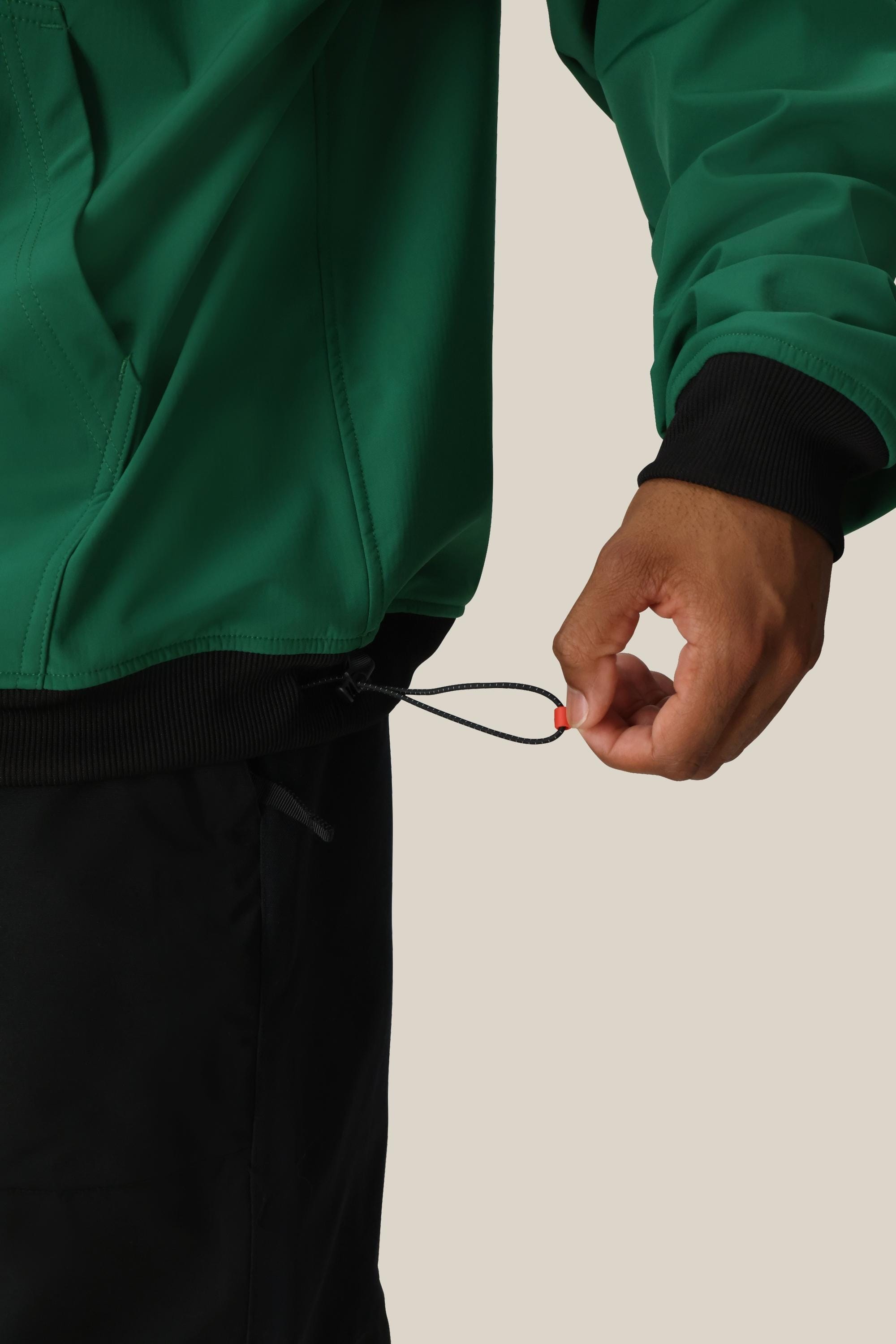 A person wearing the 686 Mens Waterproof Team Hoody in green adjusts the elastic drawstring at the hem with their right hand. They are also wearing black pants against a plain, light-colored background.