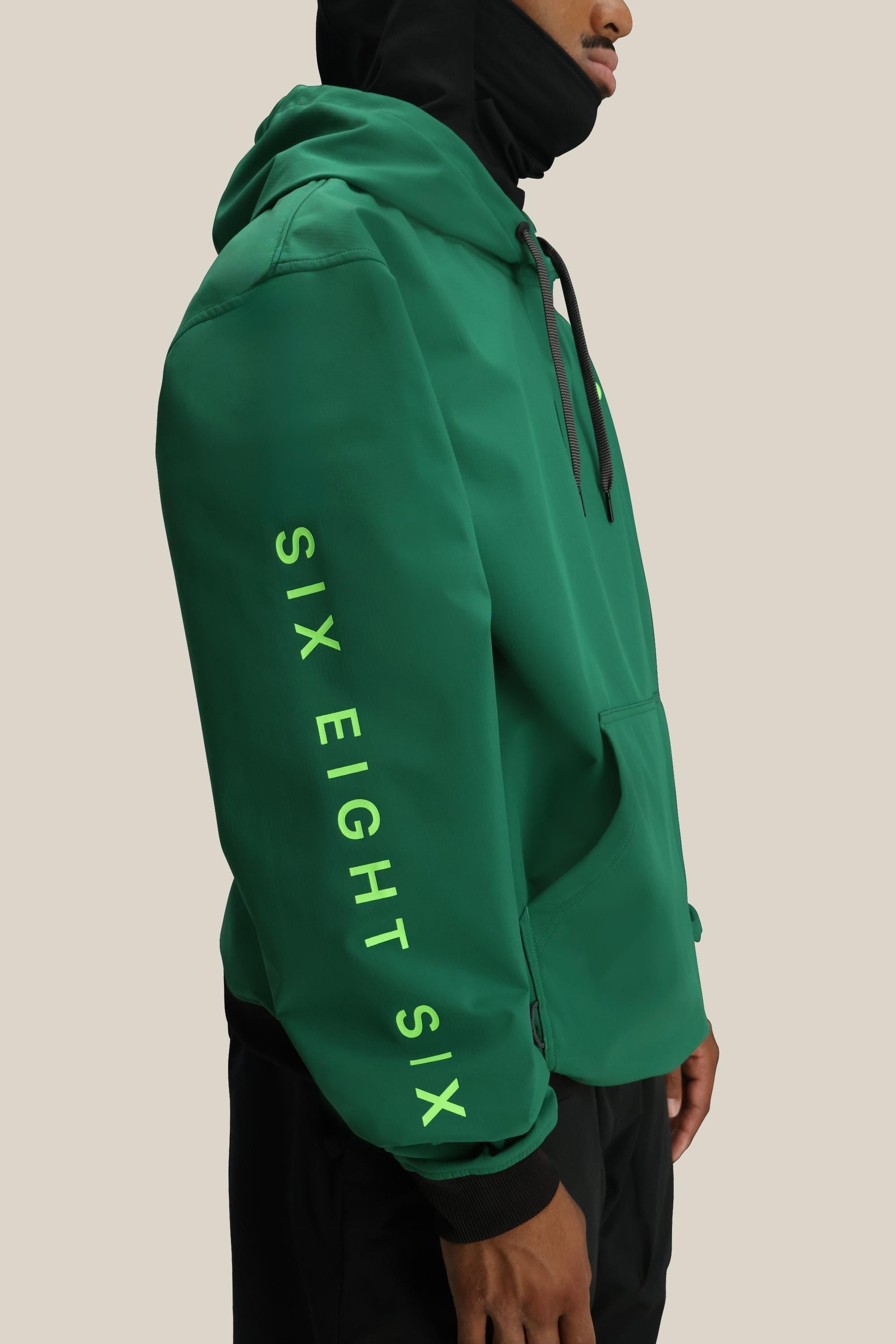 A person is wearing the 686 Mens Waterproof Team Hoody in green with black drawstrings and yellow SIX EIGHT SIX text on the sleeve, standing against a plain beige background.