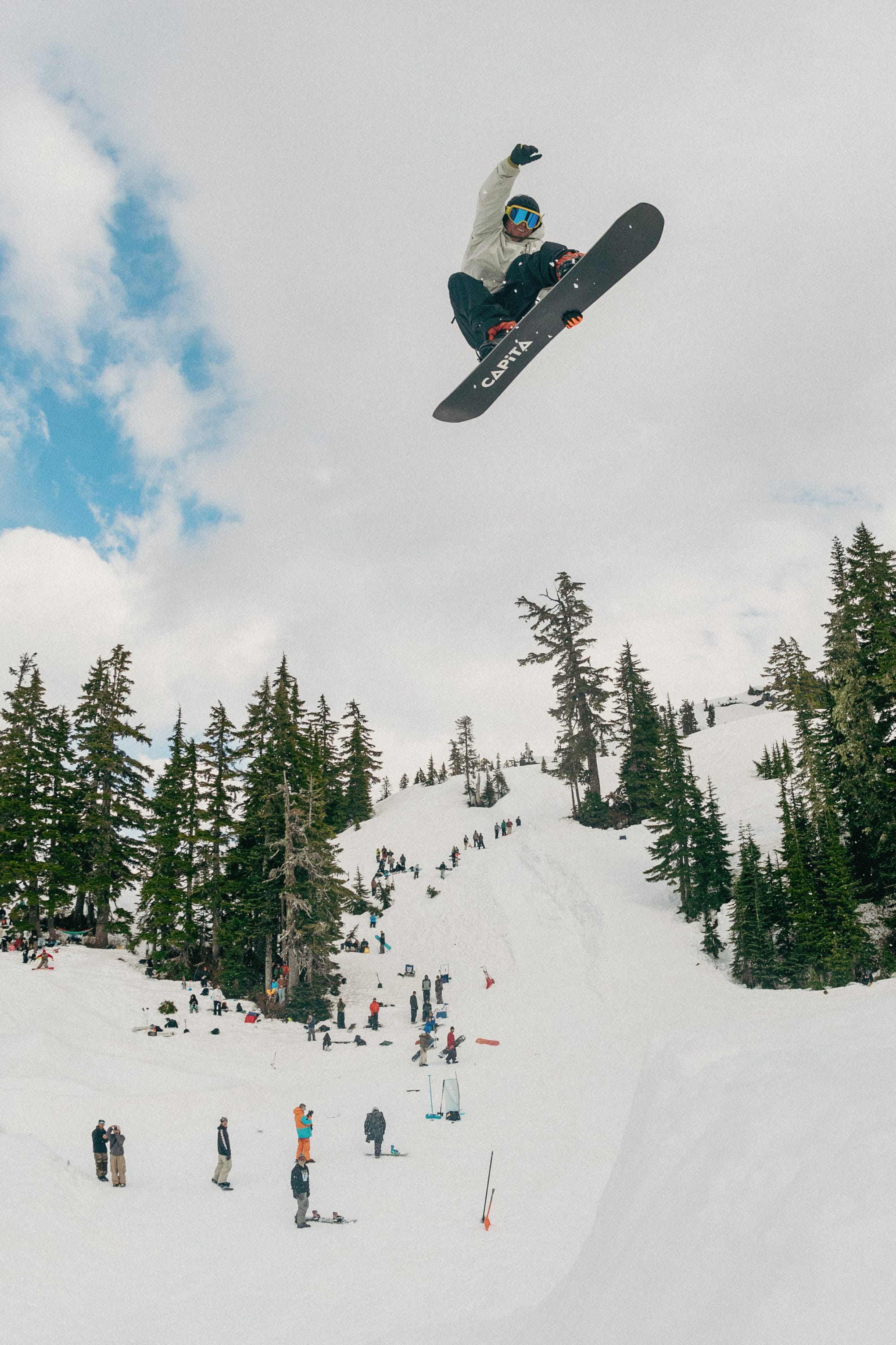 A snowboarder in the 686 Mens Waterproof Team Hoody by 686 performs a high jump above a snowy slope, with people watching below and evergreen trees in the background beneath a partly cloudy sky.
