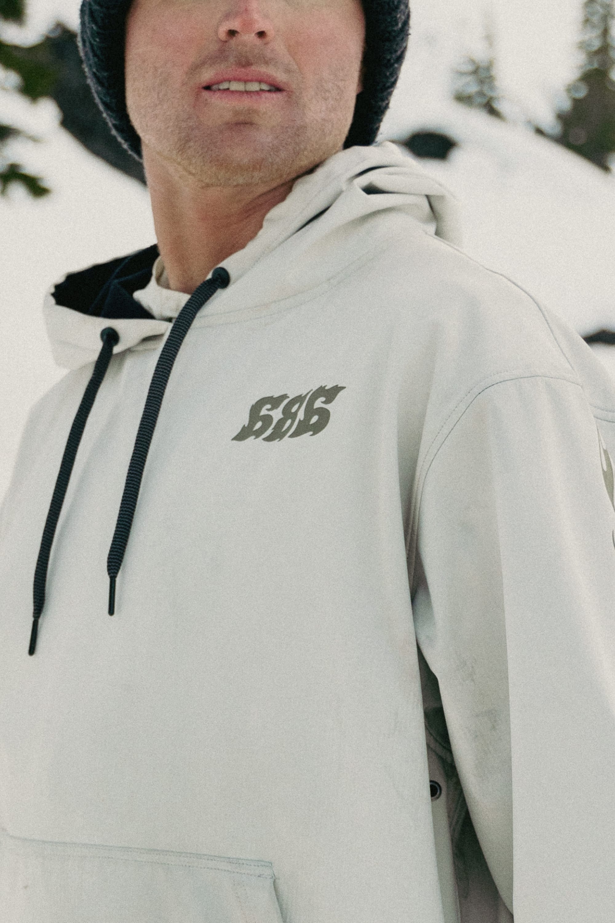 A person stands outdoors in the snow, wearing the 686 Mens Waterproof Team Hoody—a light-colored hoody with black drawstrings and the 686 logo—amid snow-covered trees.