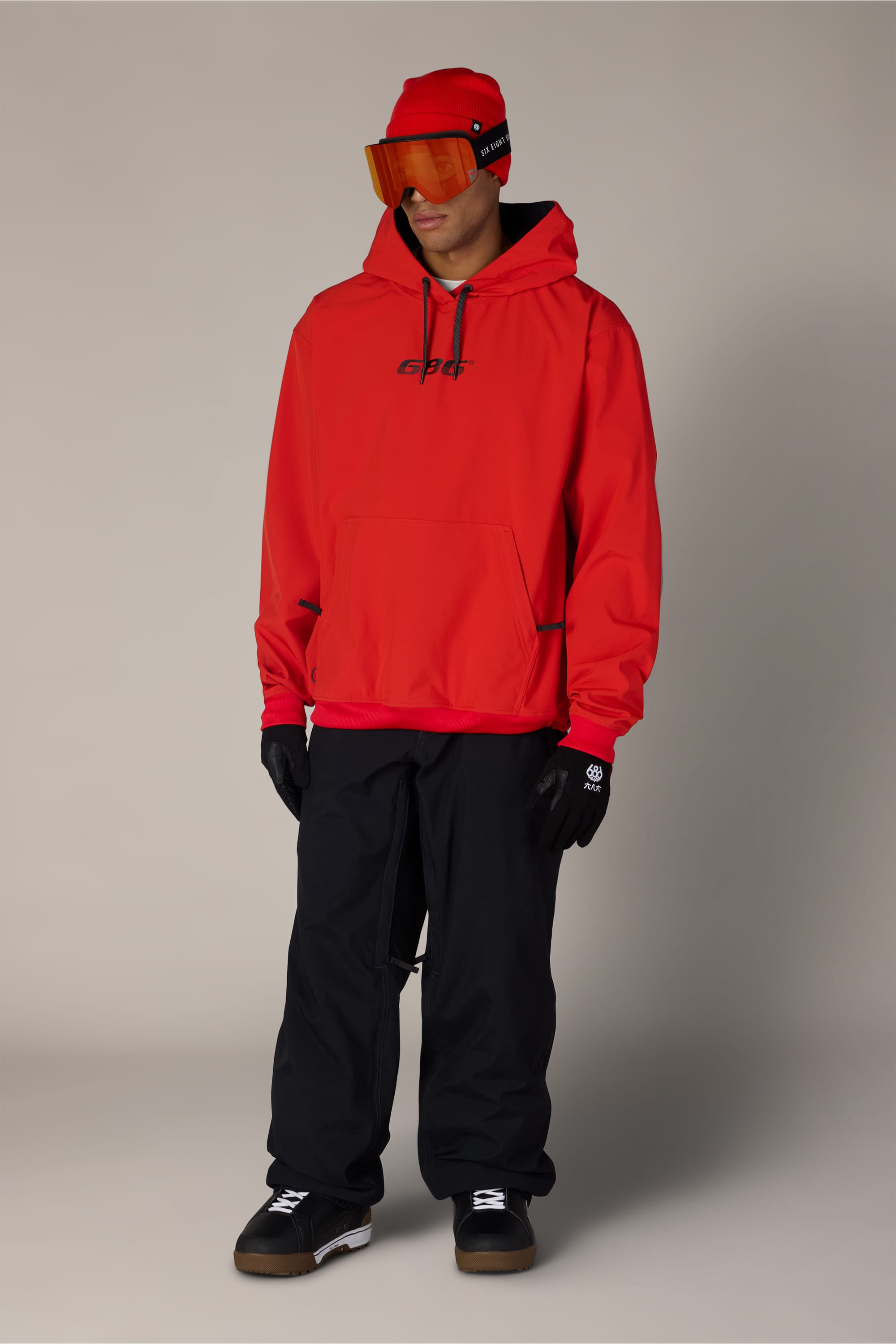 A person wearing the 686 Men’s Waterproof Team Hoody in red, black gloves, black pants, dark shoes, and an orange visor stands against a plain background.