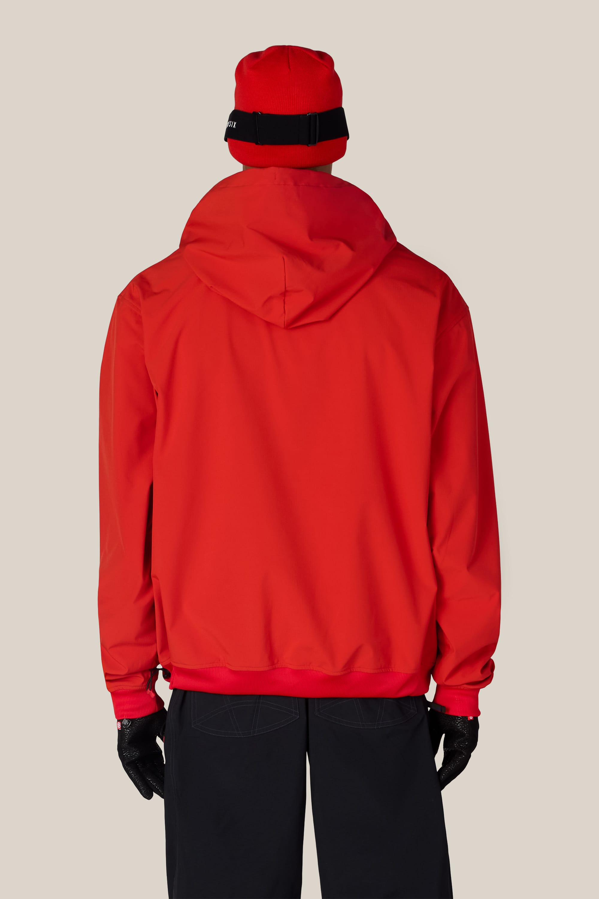 A person is standing with their back to the camera, wearing the 686 Men’s Waterproof Team Hoody in red, paired with a red beanie, black gloves, and black pants, against a plain light beige background.