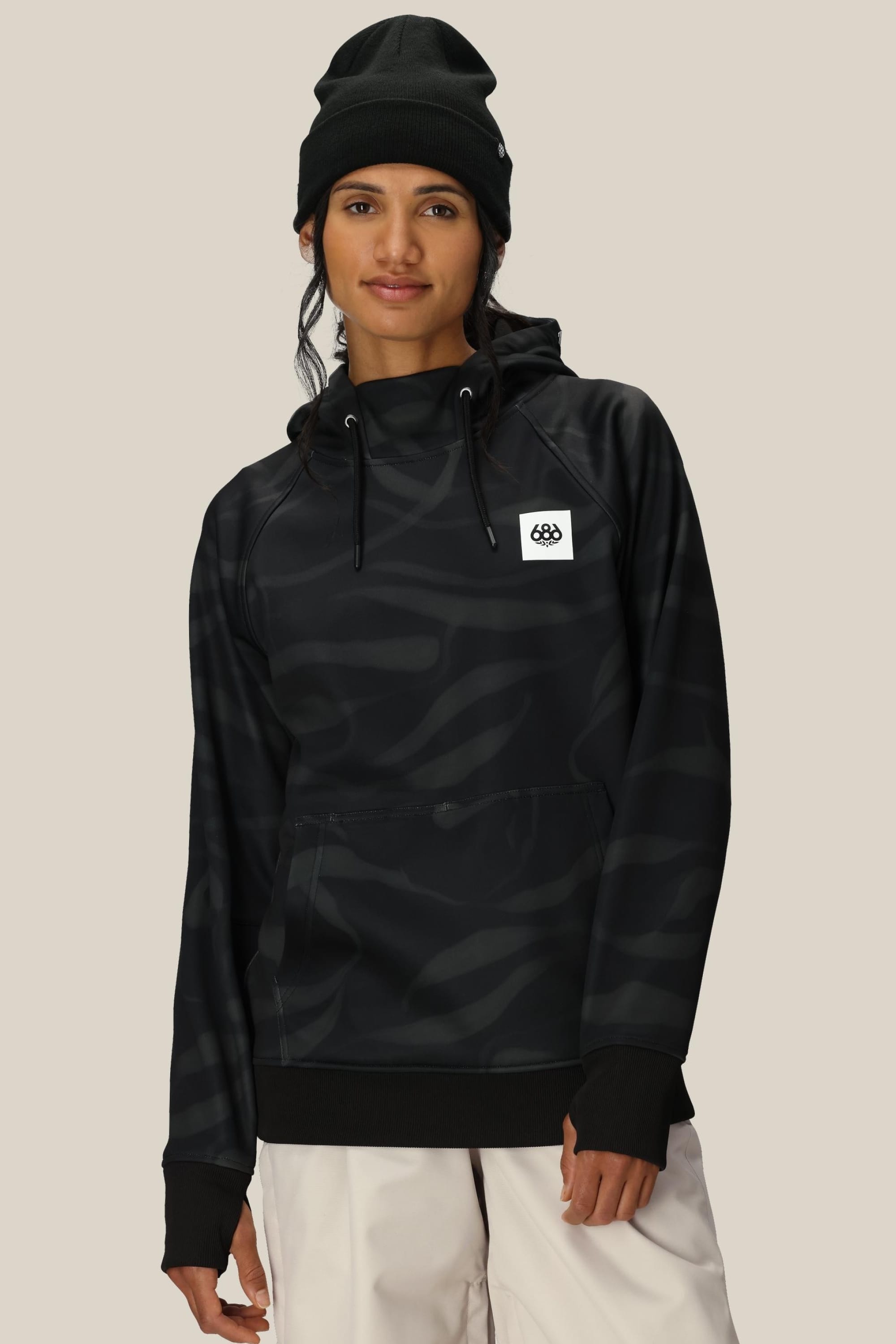 A woman wears the 686 Womens Bonded Fleece Pullover Hoody, featuring a dark pattern, fleece lining, and a white patch logo. She stands against a beige background in a black beanie, looking at the camera with a slight smile.