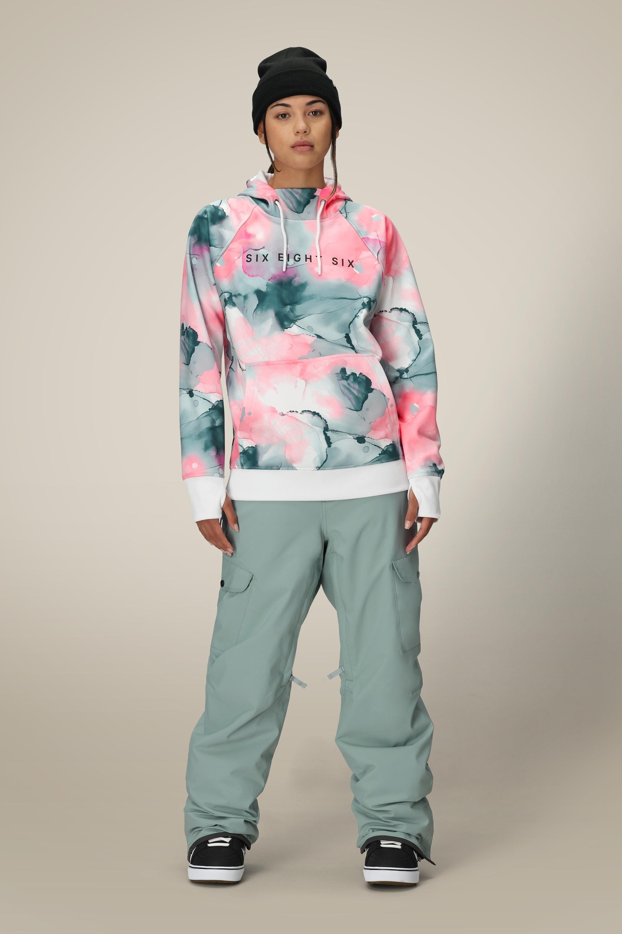 A person in a black beanie, light gray cargo pants, black-and-white sneakers, and a pink and gray floral 686 Womens Bonded Fleece Pullover Hoody stands against a neutral background.