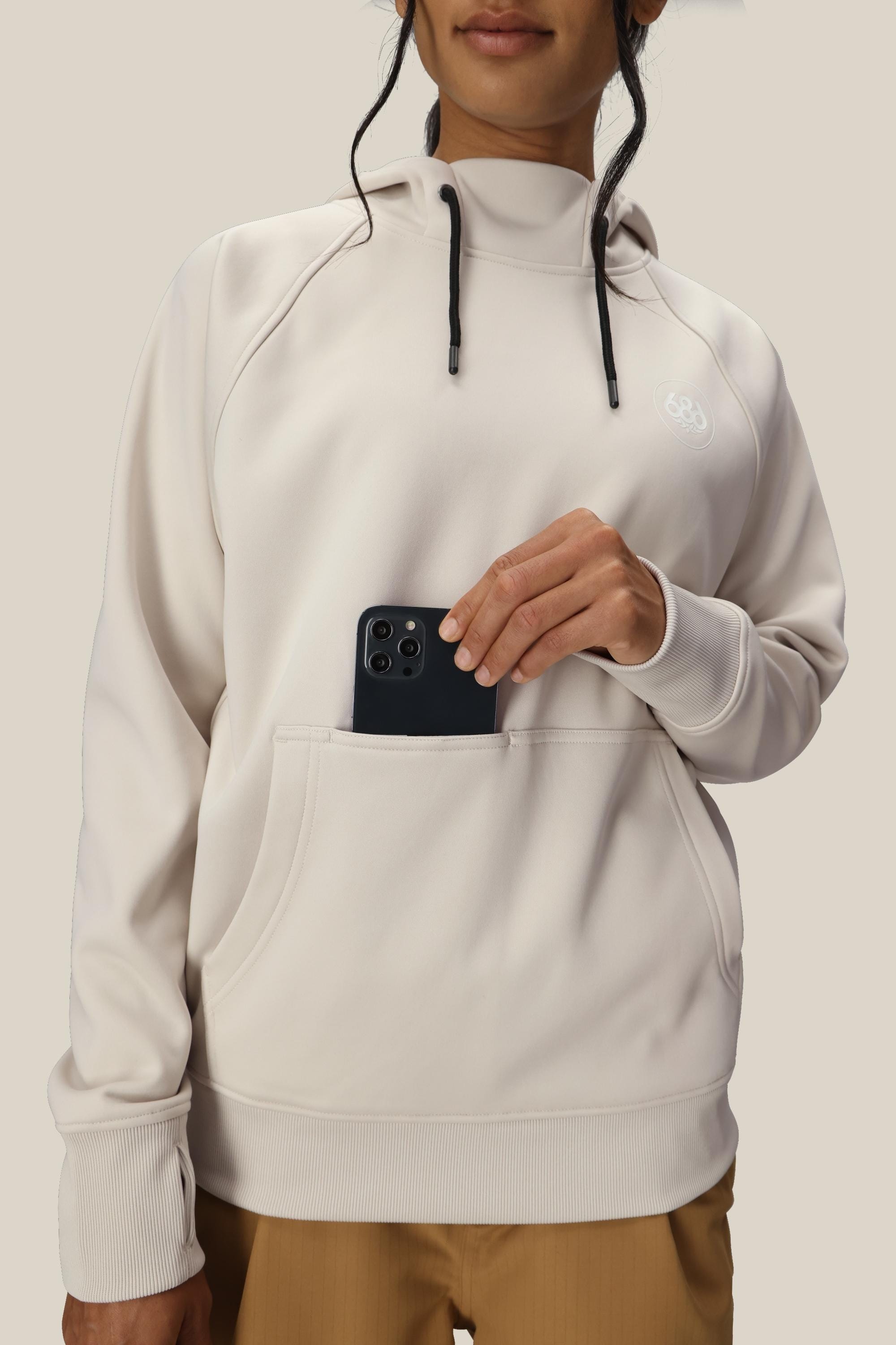 A person wearing the 686 Womens Bonded Fleece Pullover Hoody in light beige, featuring black drawstrings, a subtle chest logo, and soft fleece lining, places a black smartphone into the front pocket.