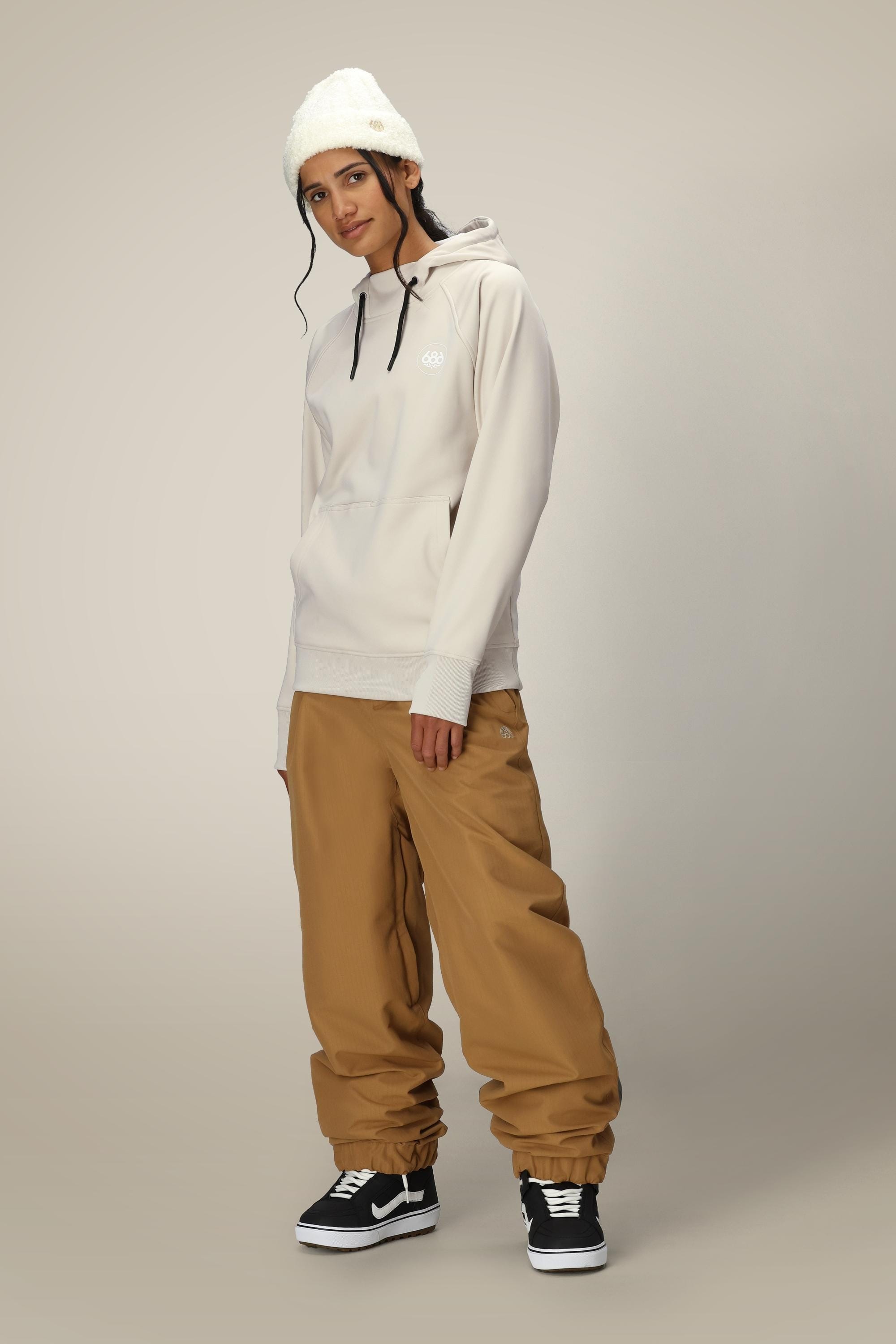 A person wearing a white beanie, the 686 Womens Bonded Fleece Pullover Hoody, loose brown pants, and black sneakers stands against a plain light background, looking at the camera with a relaxed expression.
