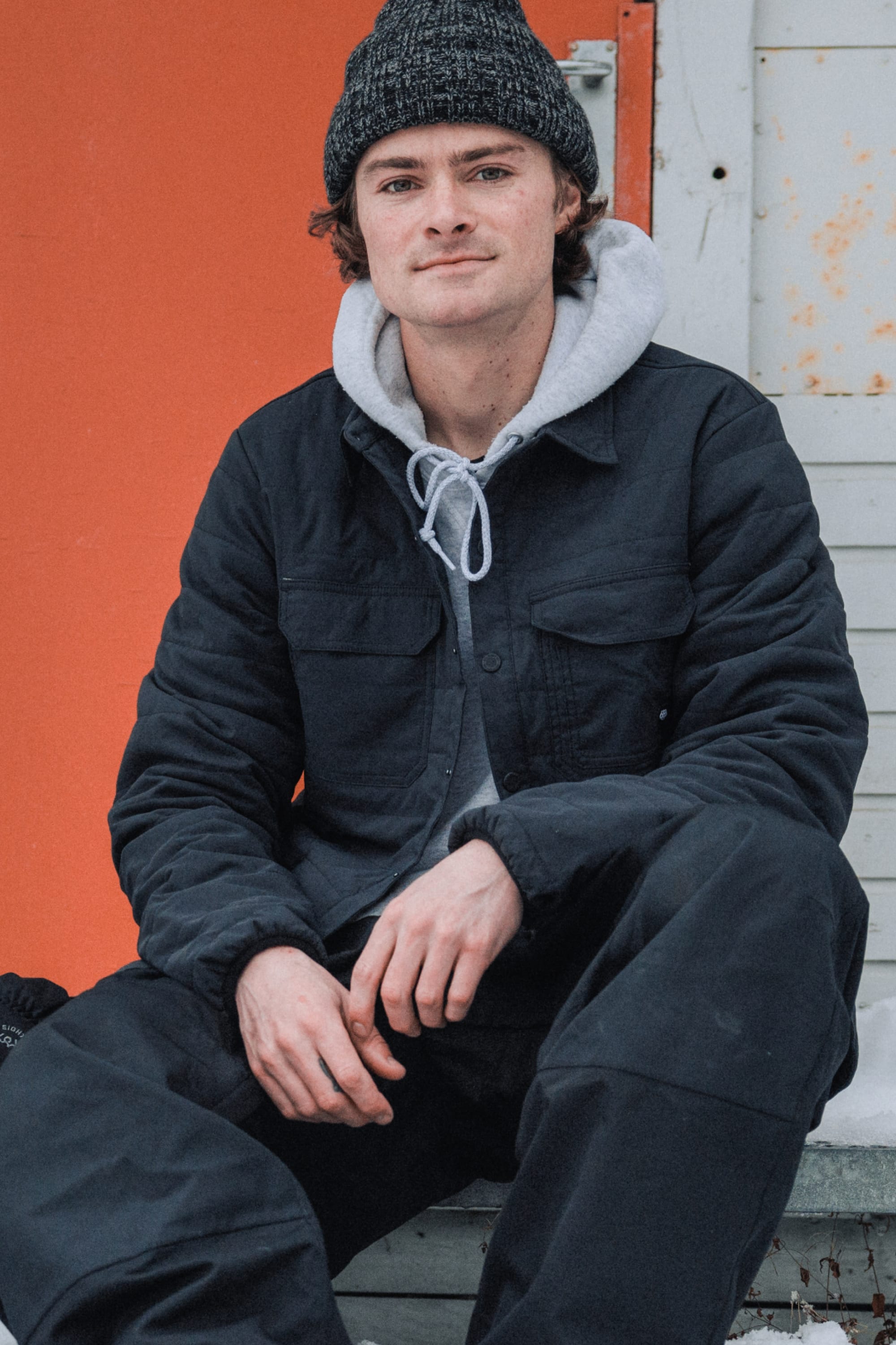A young person in stylish citywear—a gray beanie, gray hoodie, black jacket, and the 686 Men's Thermaldry™ Snap-Up Shirt—sits on outdoor steps, posing confidently before an orange and white wall.