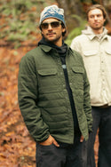 Two men are outdoors in a forest with autumn leaves. The man in front wears sunglasses, a beanie, and the 686 Men's Thermaldry™ Snap-Up Shirt—an ideal green quilted jacket for stylish citywear. The man behind him wears a beige jacket.