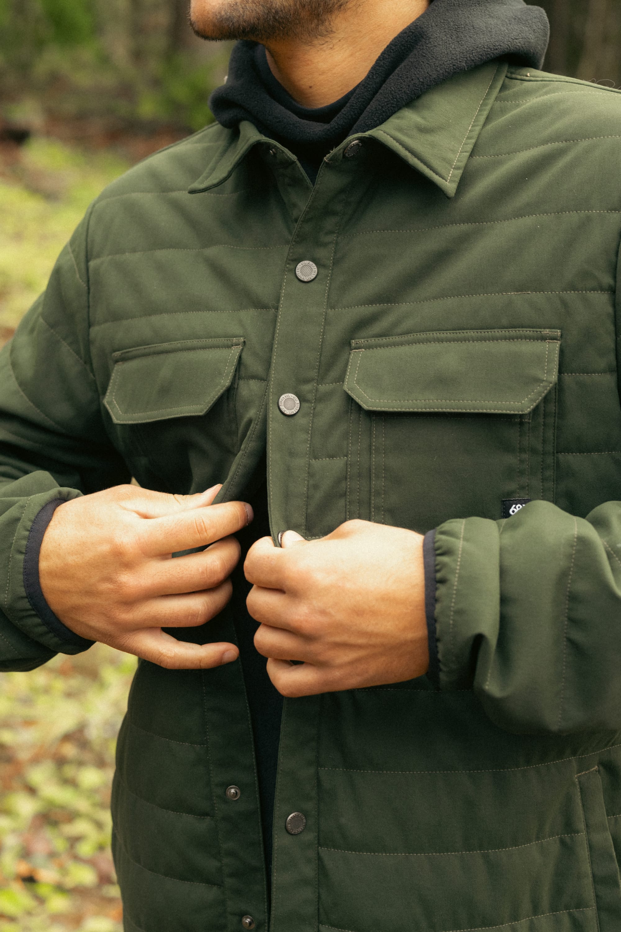 A person fastens the snaps on a 686 Men's Thermaldry™ Snap-Up Shirt, highlighting stylish citywear performance by 686. The outdoor photo focuses on the torso and hands.