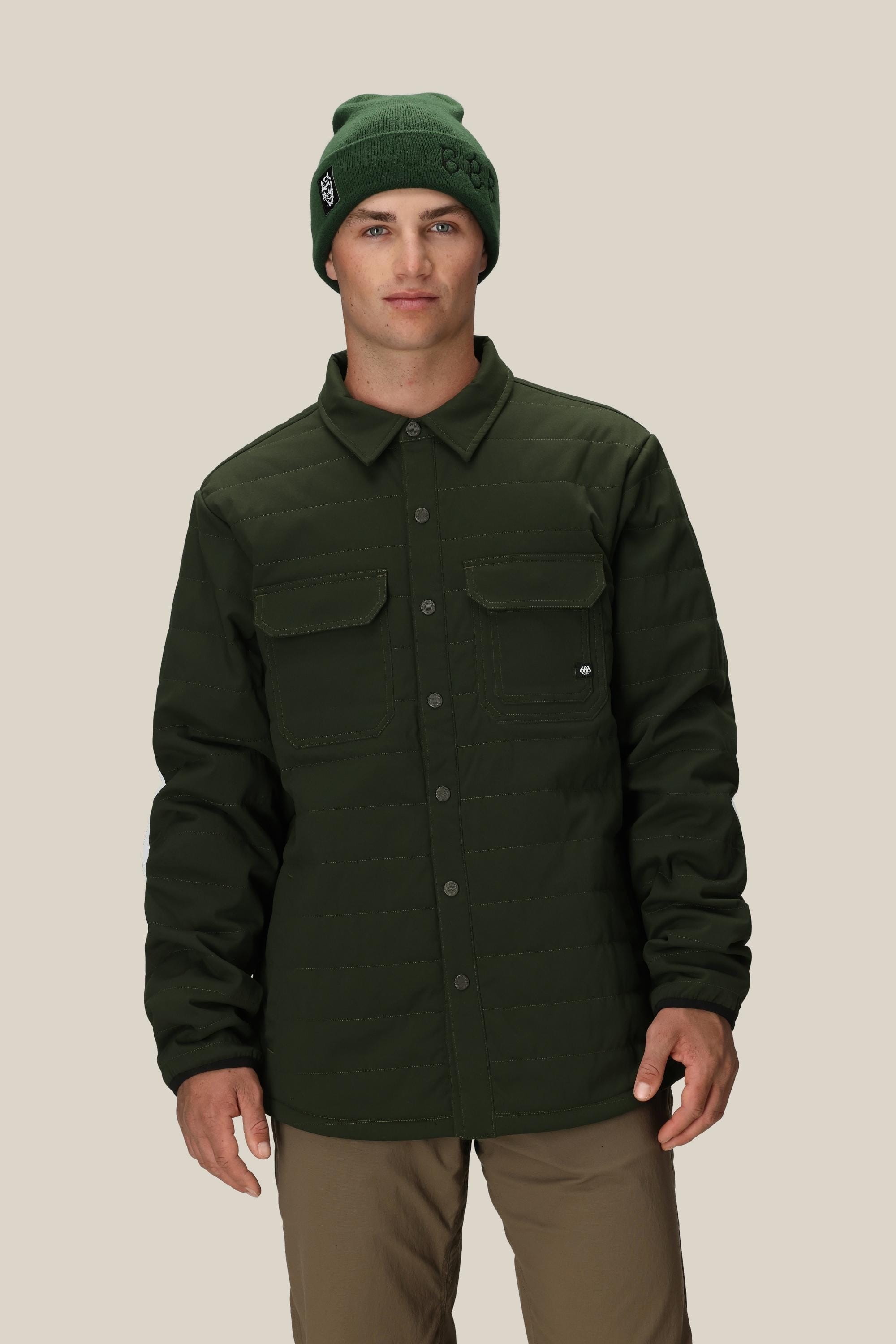 A man stands facing forward against a plain beige background, wearing the 686 Men's Thermaldry™ Snap-Up Shirt, styled with a green beanie and tan pants—an ideal look for citywear from the 686 brand.
