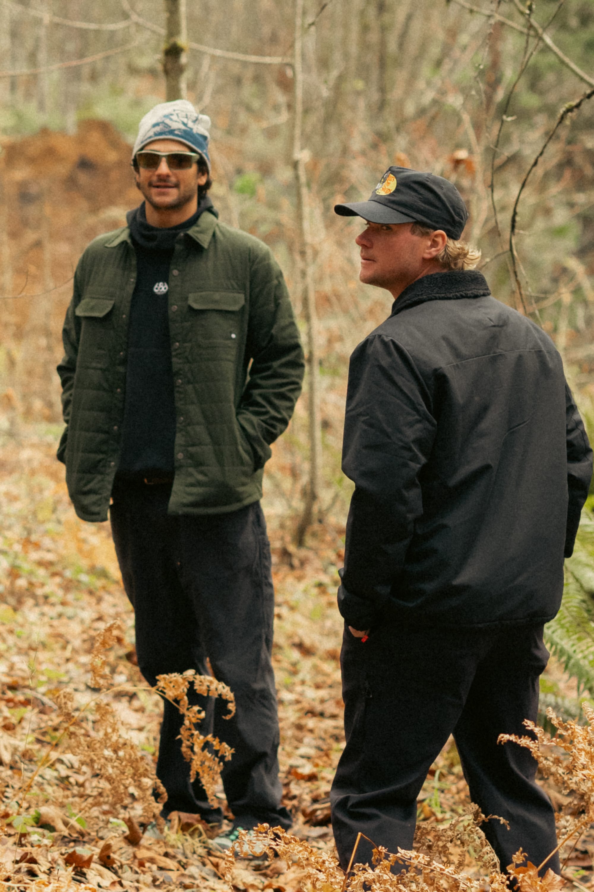 Two men stand in a forest clearing covered with dry leaves. One wears the 686 Men's Thermaldry™ Snap-Up Shirt with black pants, sunglasses, and a beanie; the other sports a black jacket, black pants, and a black cap.