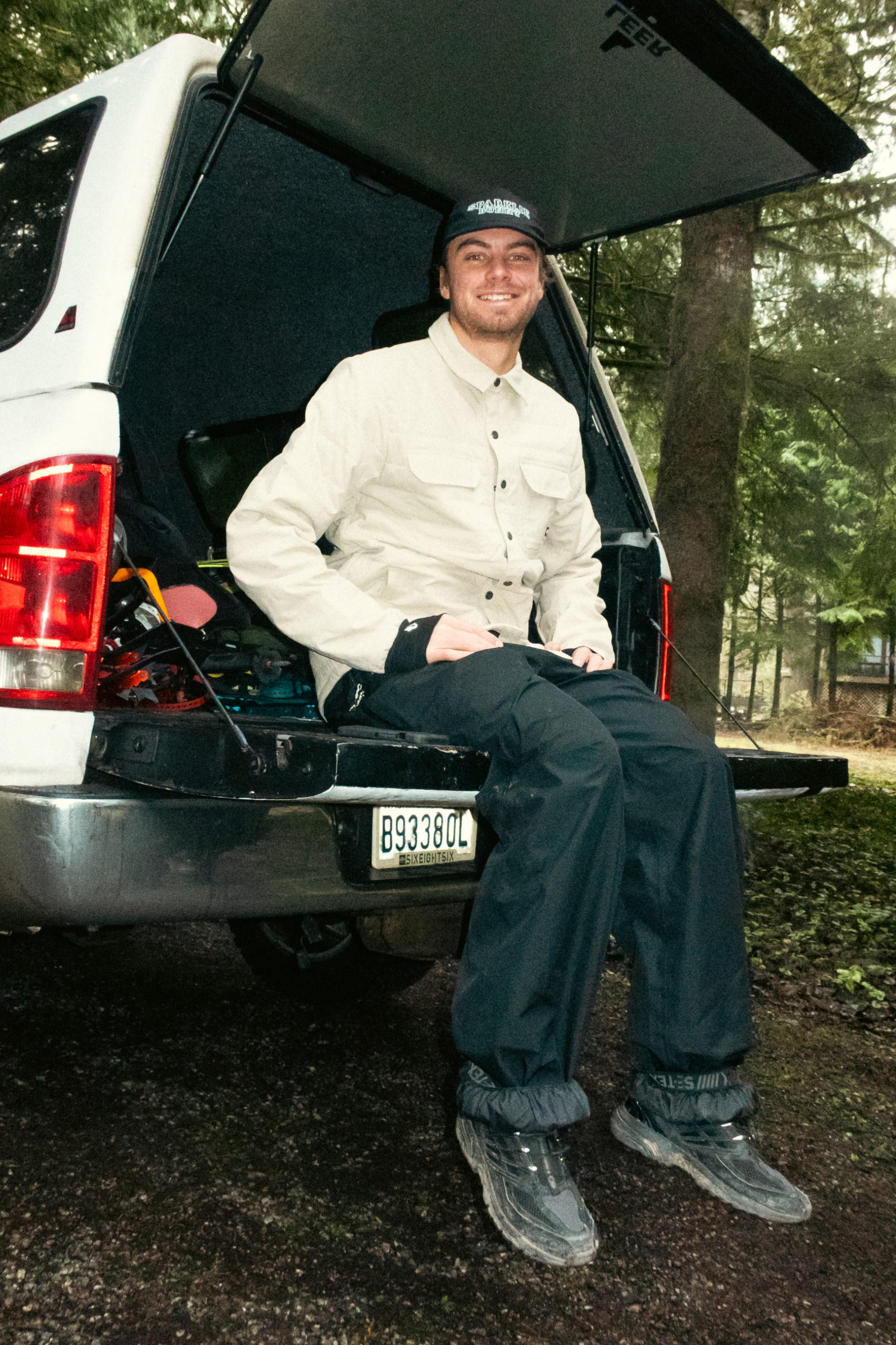 Wearing the 686 Men's Thermaldry™ Snap-Up Shirt, a man smiles from the open tailgate of a white vehicle parked in a forest, surrounded by performance gear and wet leaves on the ground.