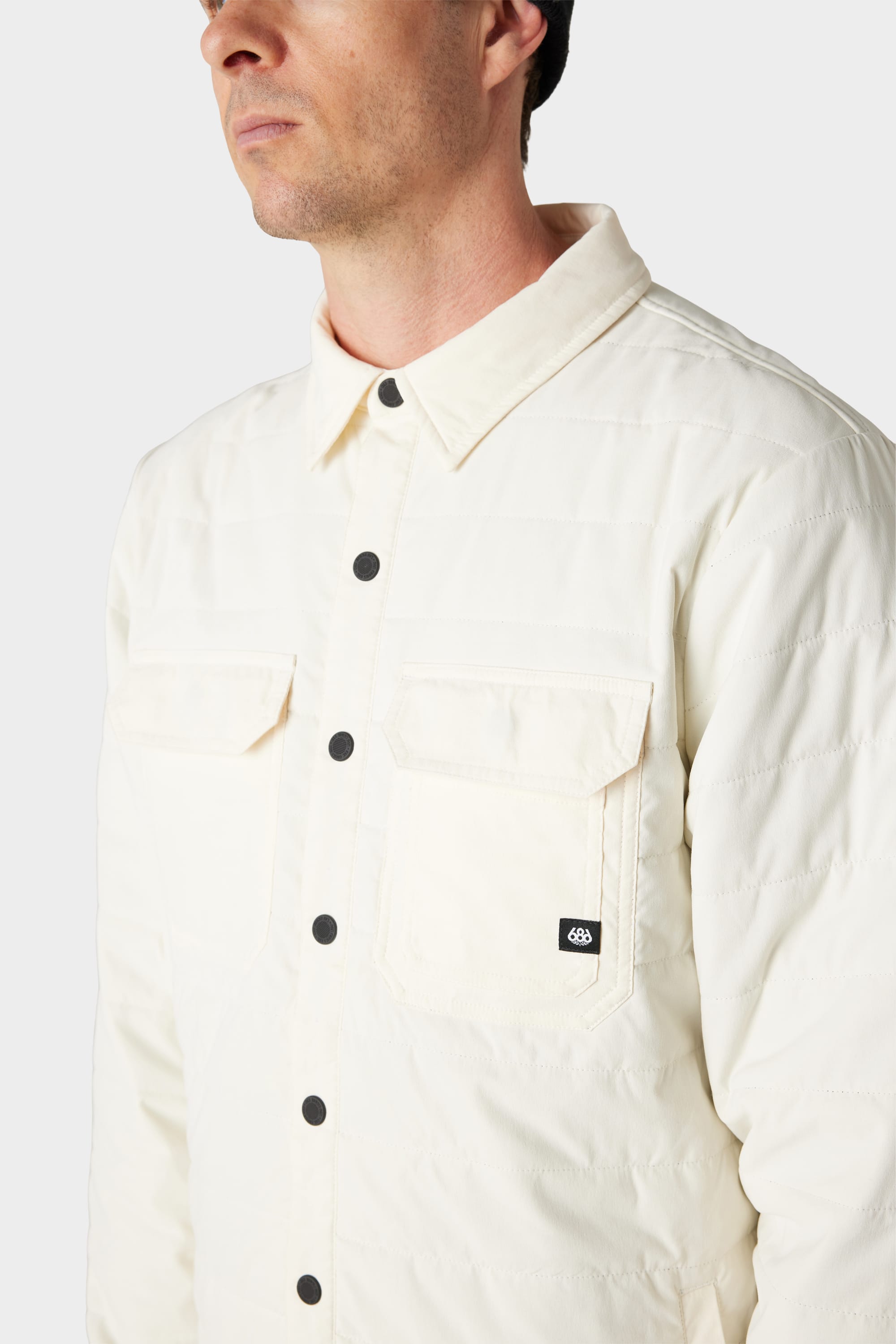 A man wears the 686 Men's Thermaldry™ Snap-Up Shirt, a cream quilted merino blend with black buttons and two chest pockets, photographed from the shoulders up against a white background, highlighting 686's stylish citywear.
