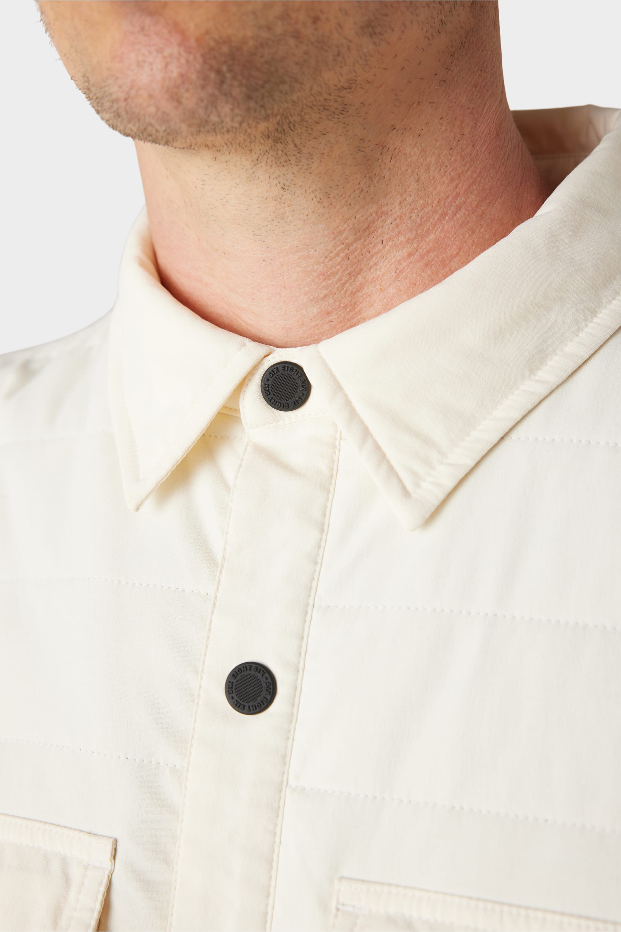 A close-up of a person wearing the 686 Men's Thermaldry™ Snap-Up Shirt, a cream-colored merino blend with black snaps. The image highlights the shirt’s collar, two snaps, and part of the neck—ideal for stylish citywear.