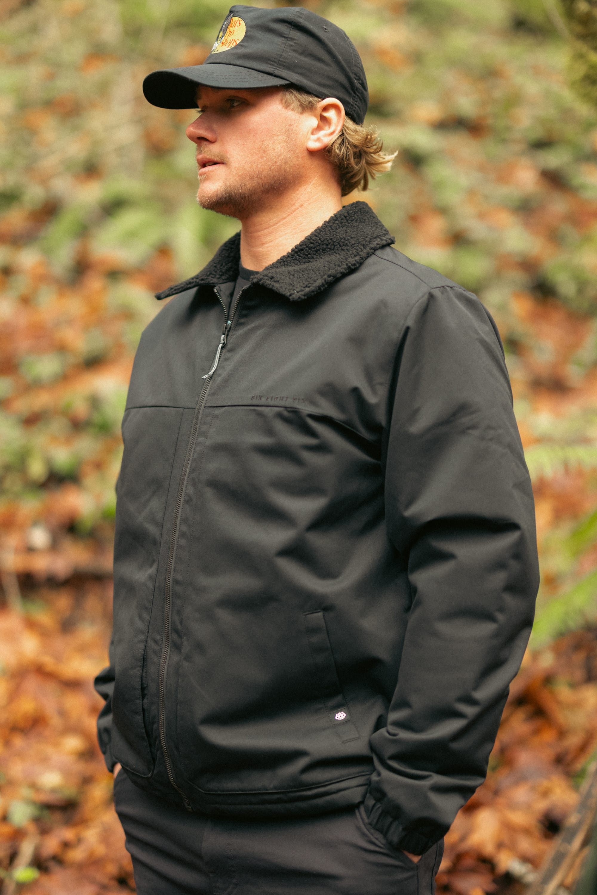 A person wearing the 686 Mens Caim Insulated Jacket and a black cap stands outdoors in a forest, surrounded by fallen leaves and greenery.