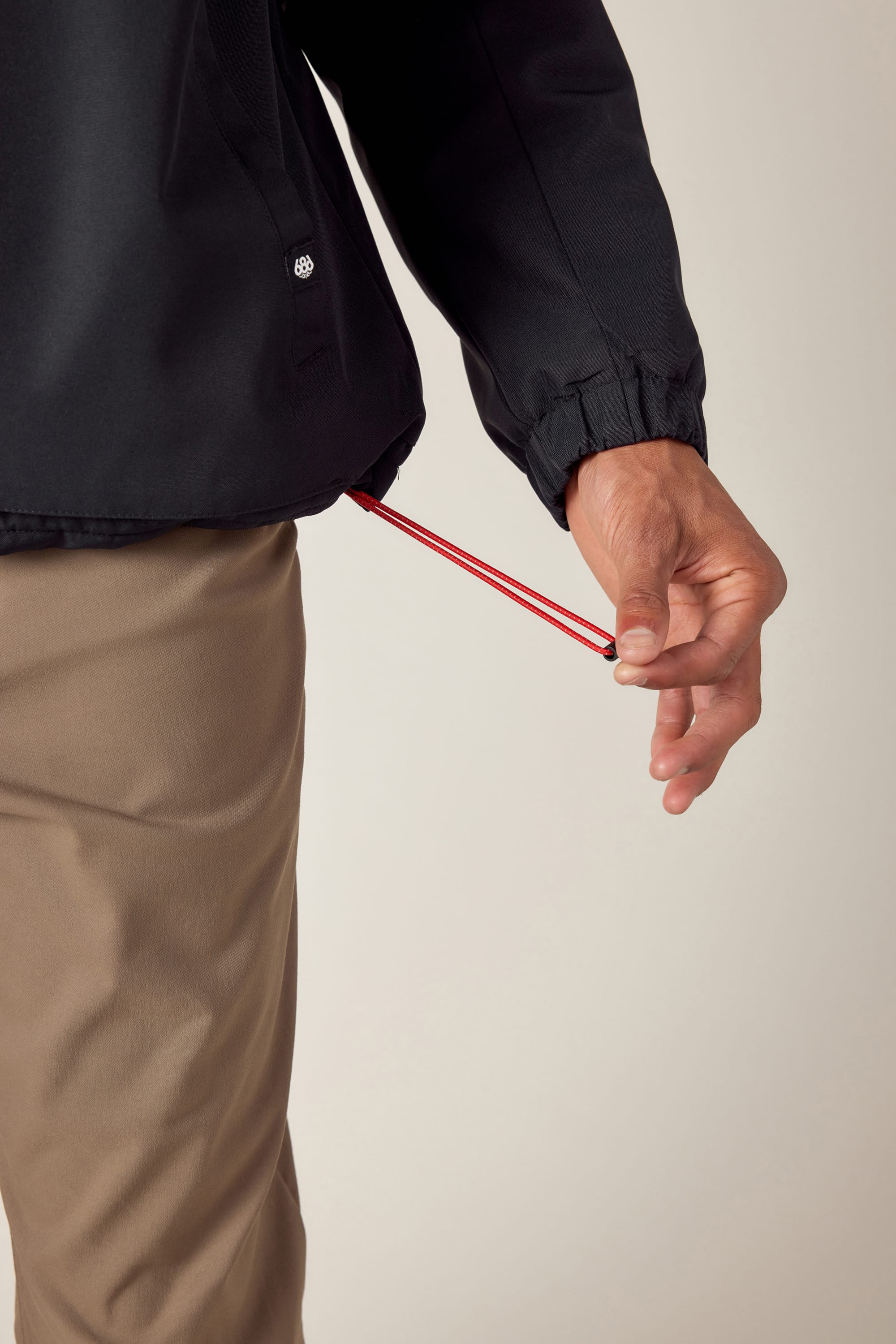 A person adjusts the red hem drawstring on a 686 Mens Caim Insulated Jacket in black, paired with tan pants. The lower torso, arm, and hand are shown against a plain background.