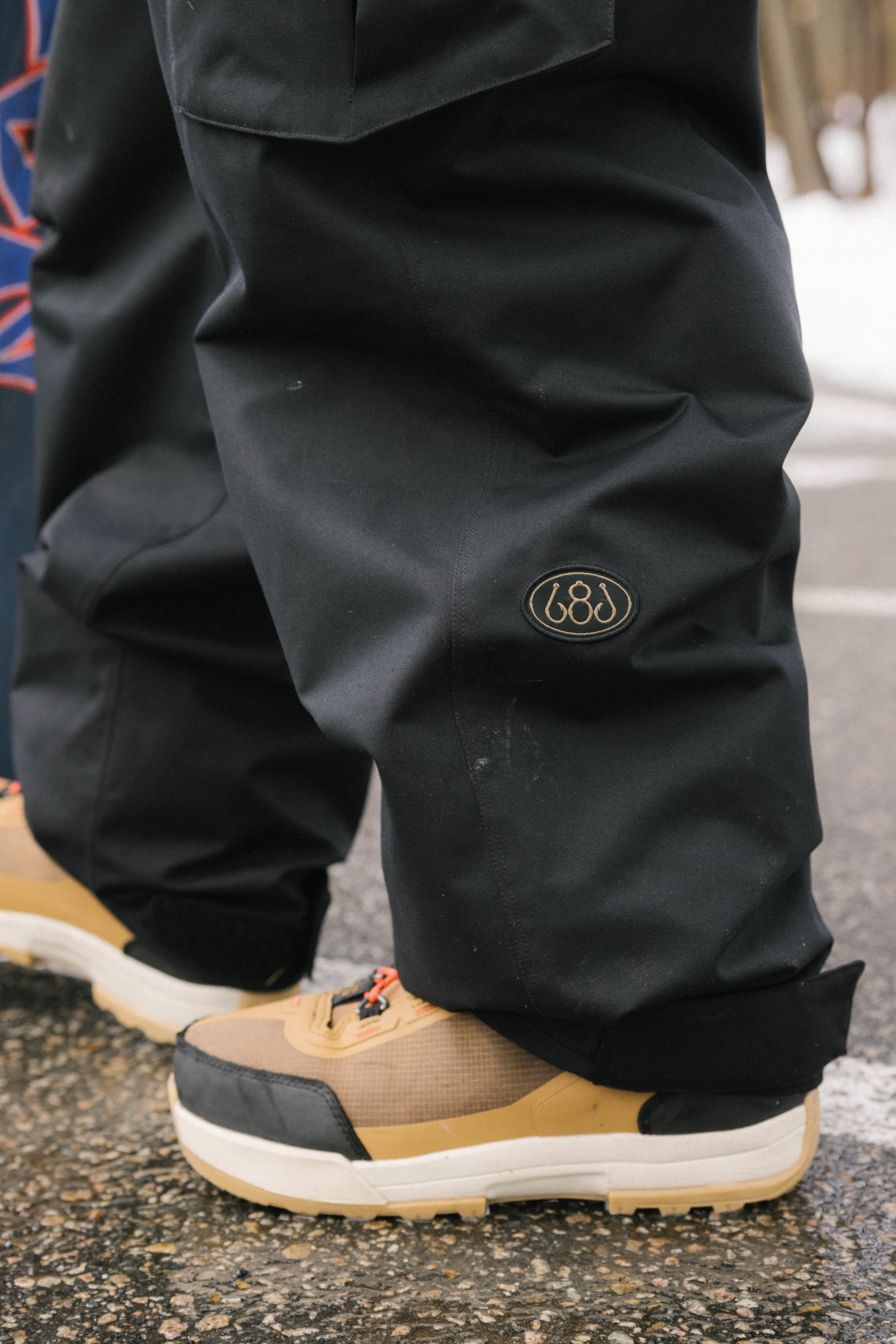 Close-up of a person wearing 686 Men's Essox™ Cargo Pant—black, baggy with cuff adjusters and a round "686" patch—paired with beige sneakers featuring black and white soles, standing on a wet surface.