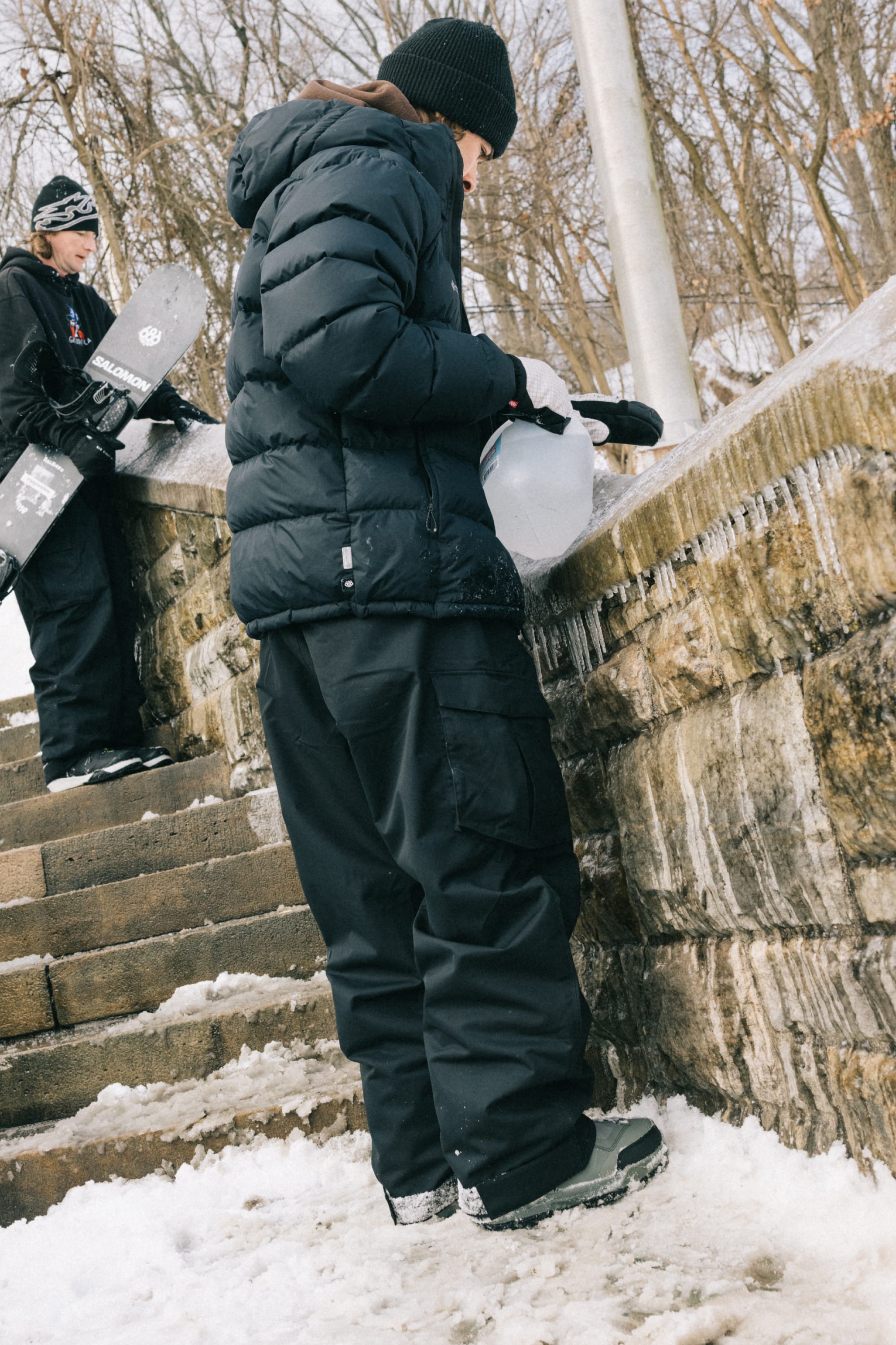 Two people dressed for winter stand on snowy steps beside a stone wall—one pours liquid from a jug onto the stone while the other, wearing the 686 Men’s Essox™ Cargo Pant by 686, holds a snowboard behind them. Bare trees and deep snow fill the wintry background.