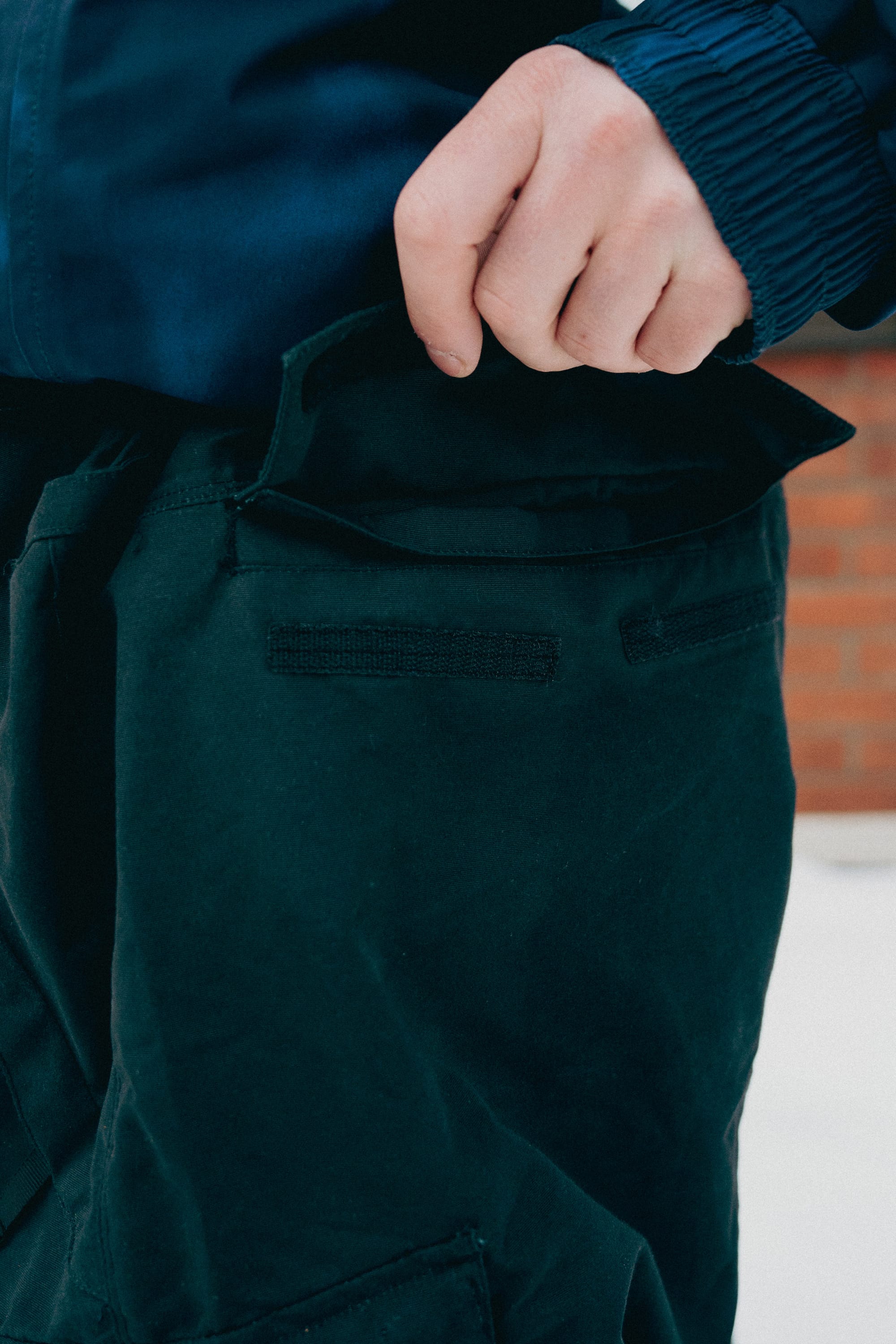 A person wearing 686 Men's Essox™ Cargo Pant in dark green with cuff adjusters holds open a front pocket, with a brick wall blurred in the background.