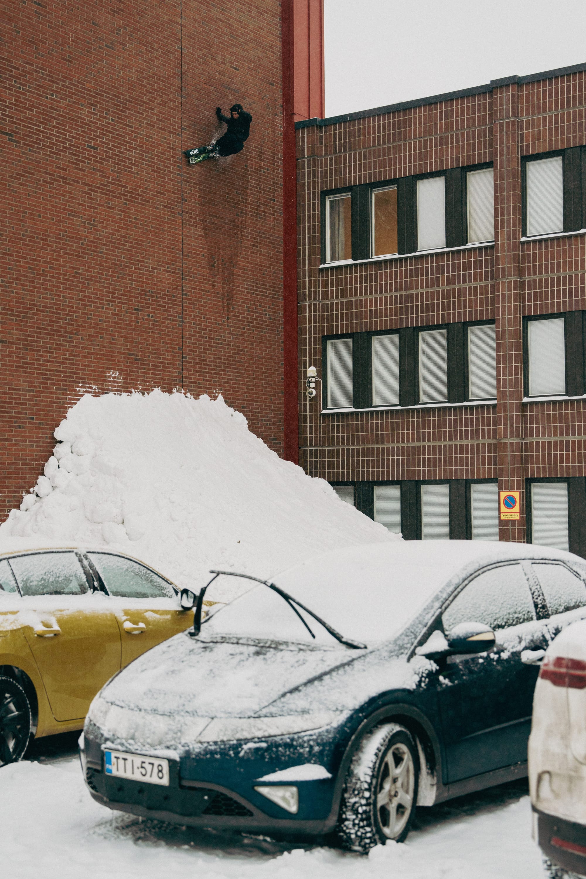 Wearing 686 Men's Essox™ Cargo Pants, a snowboarder rides up a snow ramp and vertically up a red brick wall in a snowy parking lot, with parked cars covered in snow visible in the foreground.