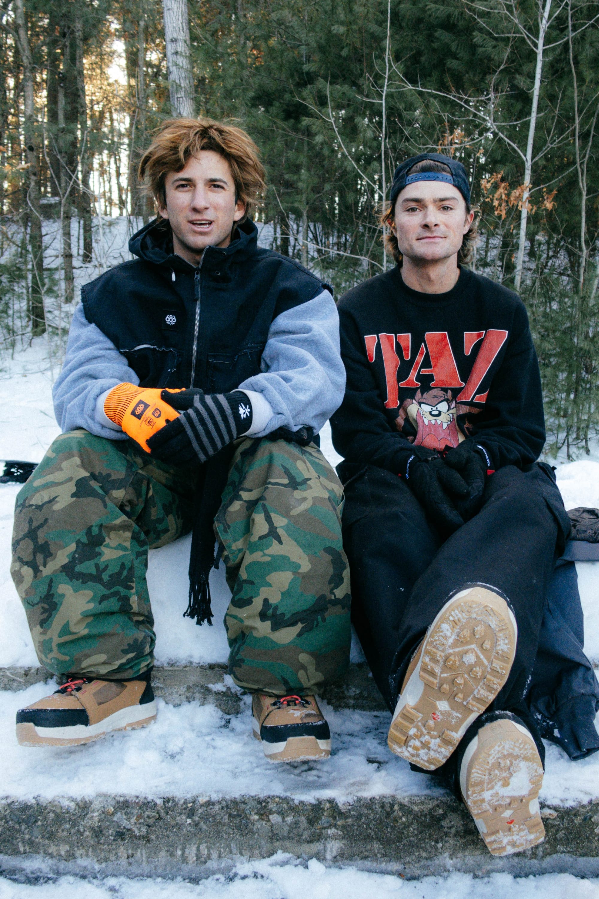 Two young men in winter gear sit on a snowy forest ledge. One wears a black jacket and 686 Men's Essox™ Cargo Pant, holding orange gloves; the other sports a black cap, Taz graphic sweater, and displays his boot soles.