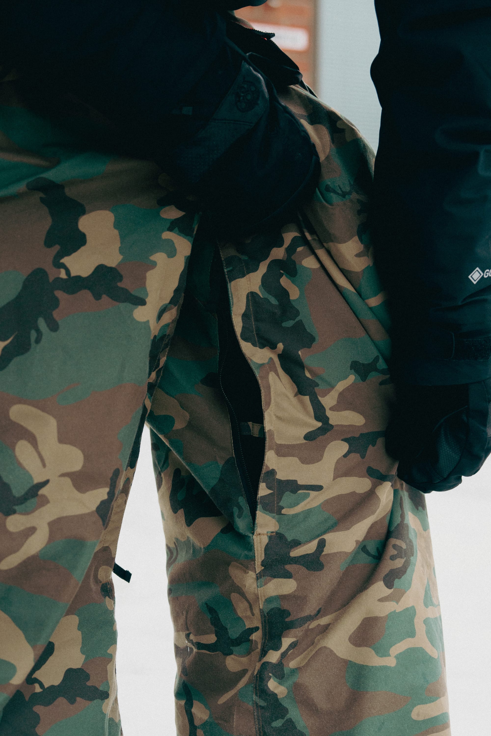 A person wearing 686 Men's Essox™ Cargo Pant by 686 with black gloves displays a large seam tear revealing the inner fabric, while snow covers the ground in the background.