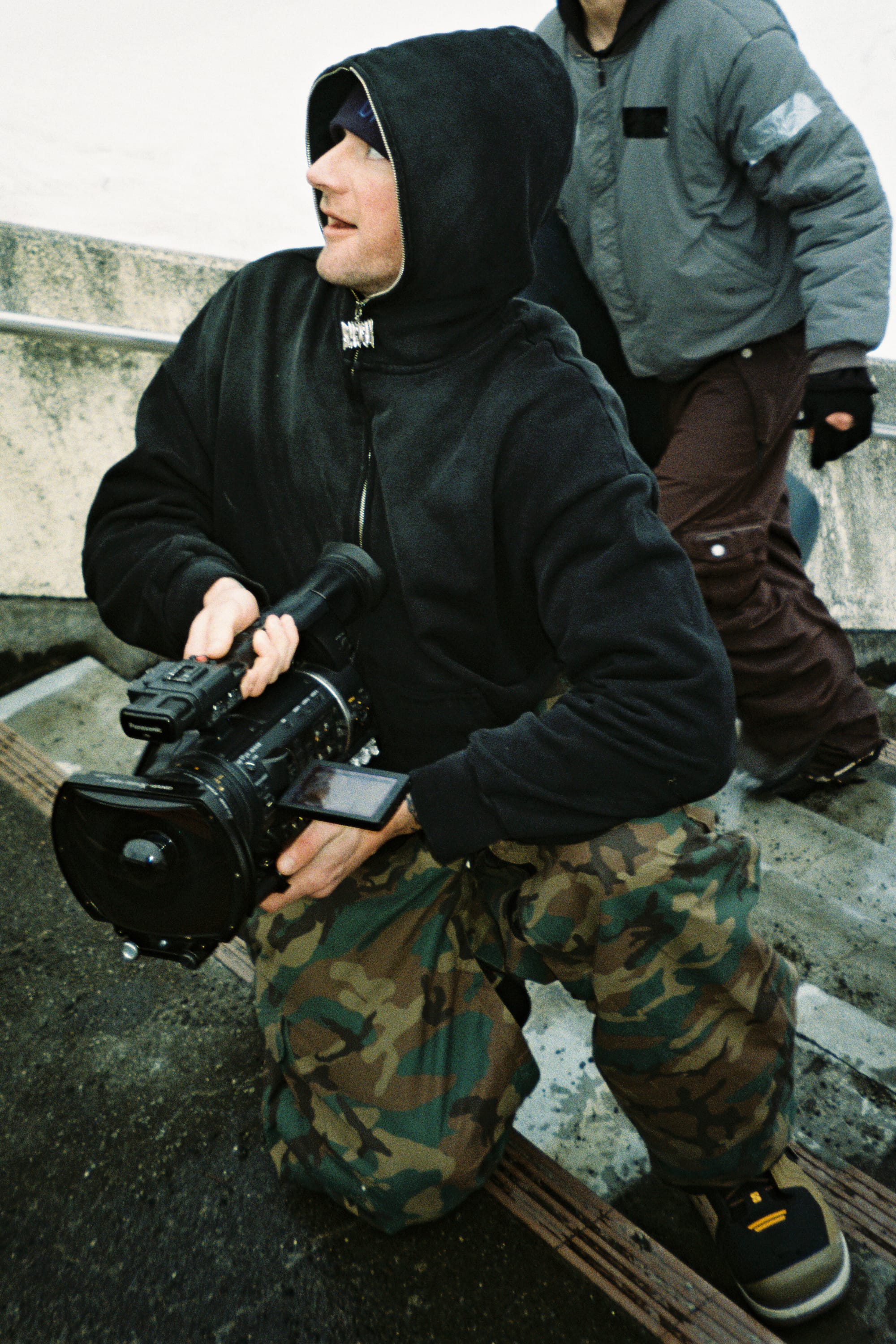 A person wearing a black hoodie and 686 Men’s Essox™ Cargo Pant with cuff adjusters kneels on the ground holding a large video camera, while another in a grey jacket stands behind them outdoors by a concrete structure.