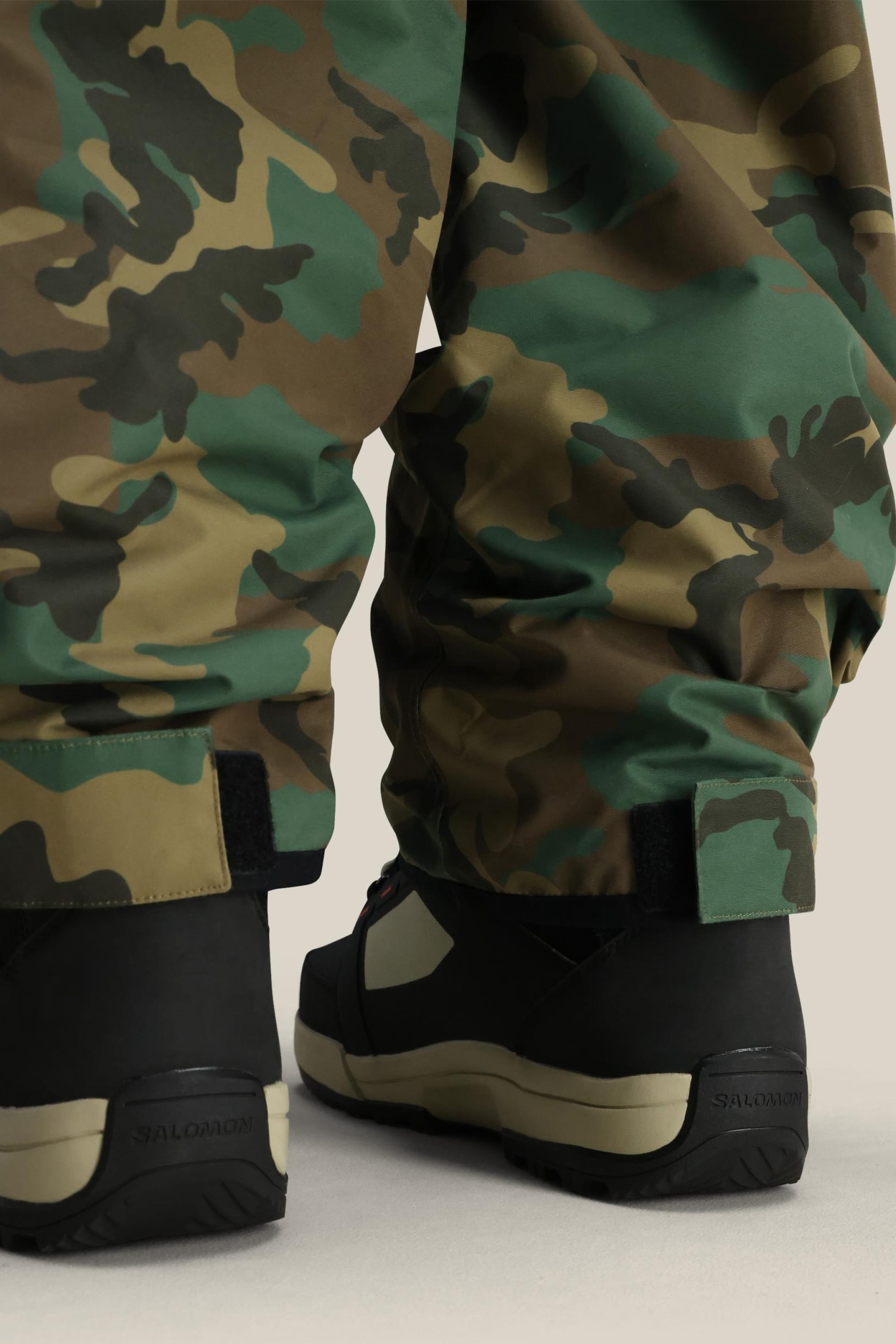 A rear close-up of a person’s lower legs wearing 686 Men's Essox™ Cargo Pants in green camo, featuring cuff adjusters, paired with black Salomon hiking boots.