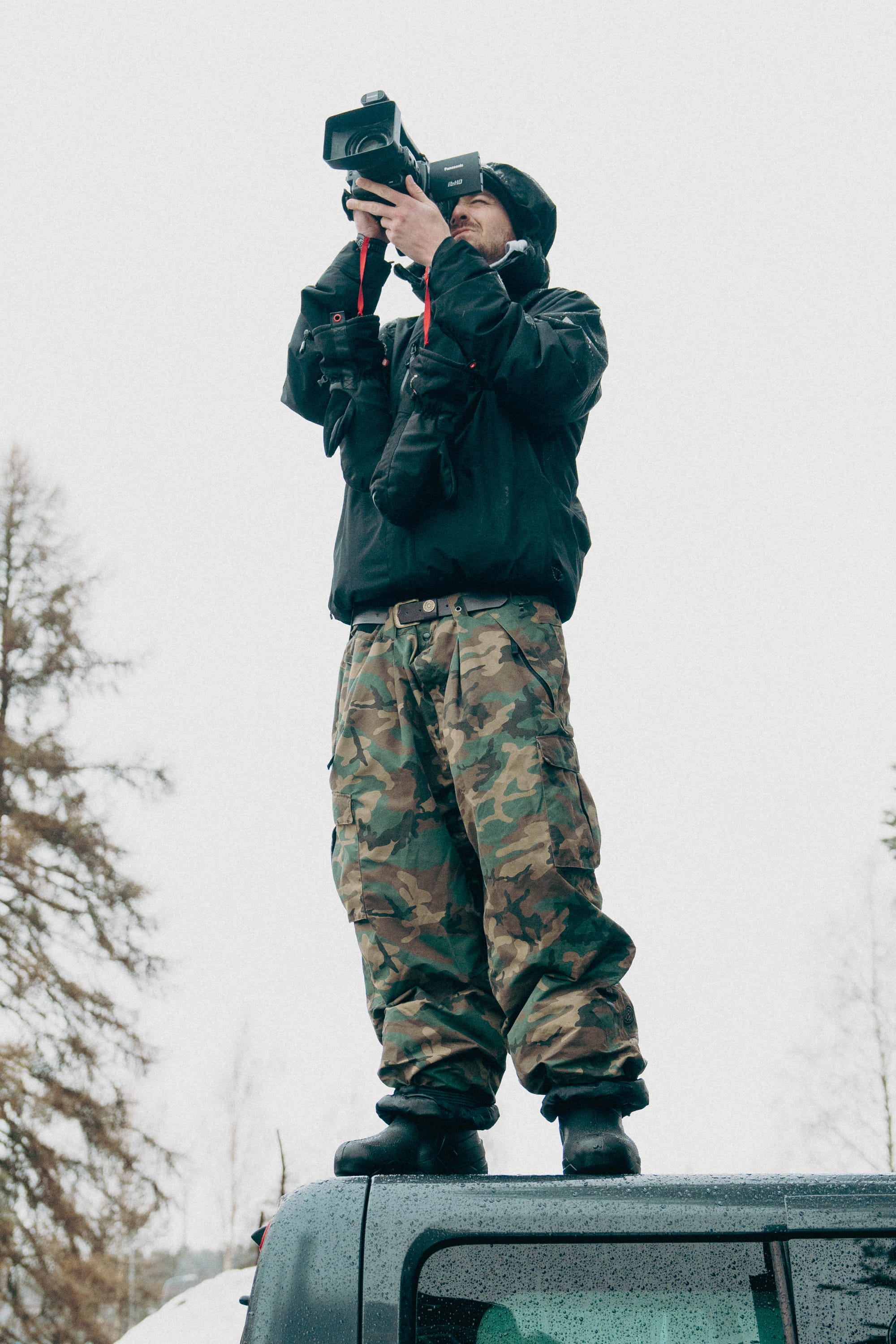 Wearing 686 Men's Essox™ Cargo Pant from 686 and a black jacket, a person stands atop a vehicle filming with a large video camera. Bare trees are seen in the background under an overcast sky.