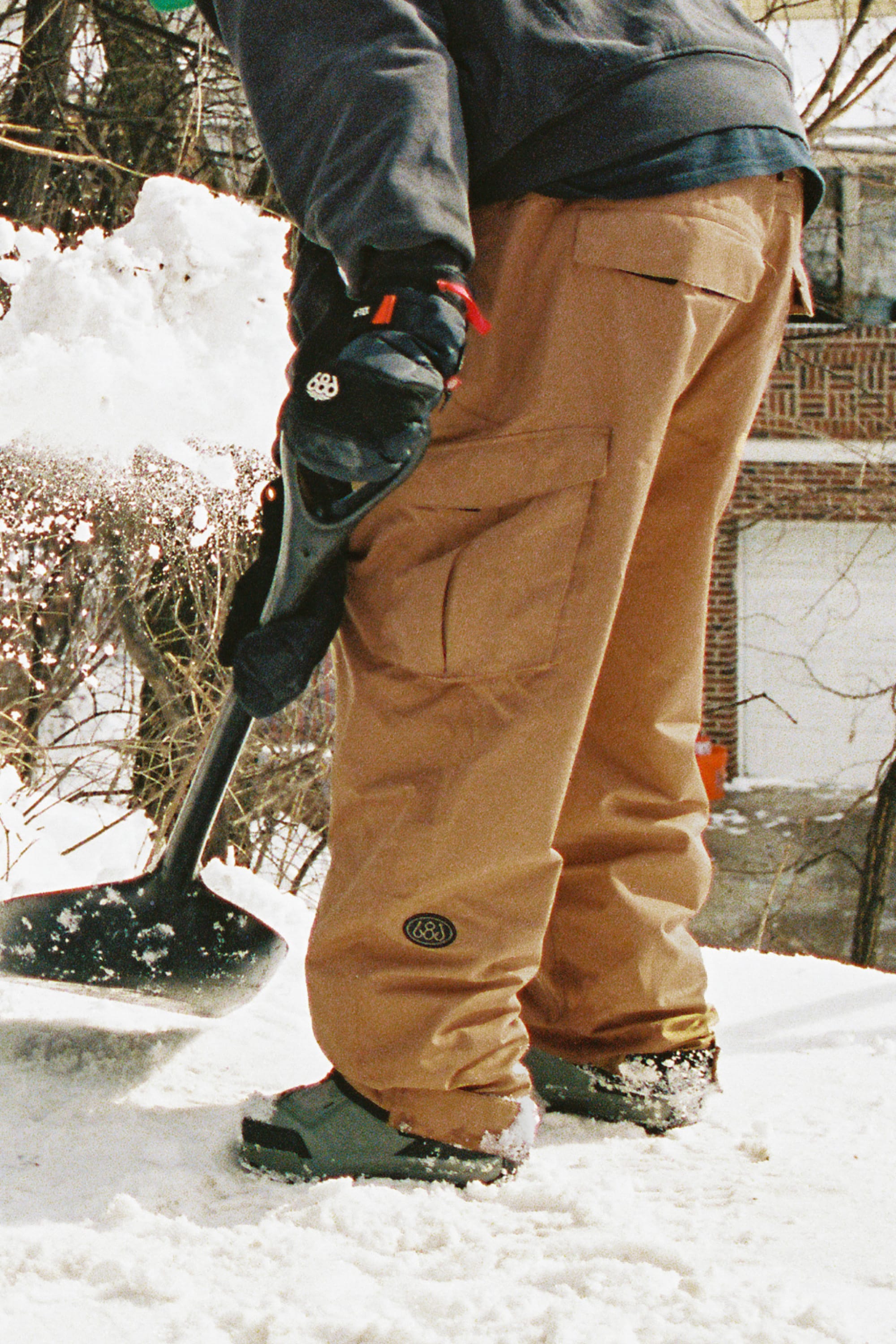 A person dressed in 686 Men’s Essox™ Cargo Pant and black gloves shovels snow outside, sending snow flying from the shovel, with a brick building visible in the background.